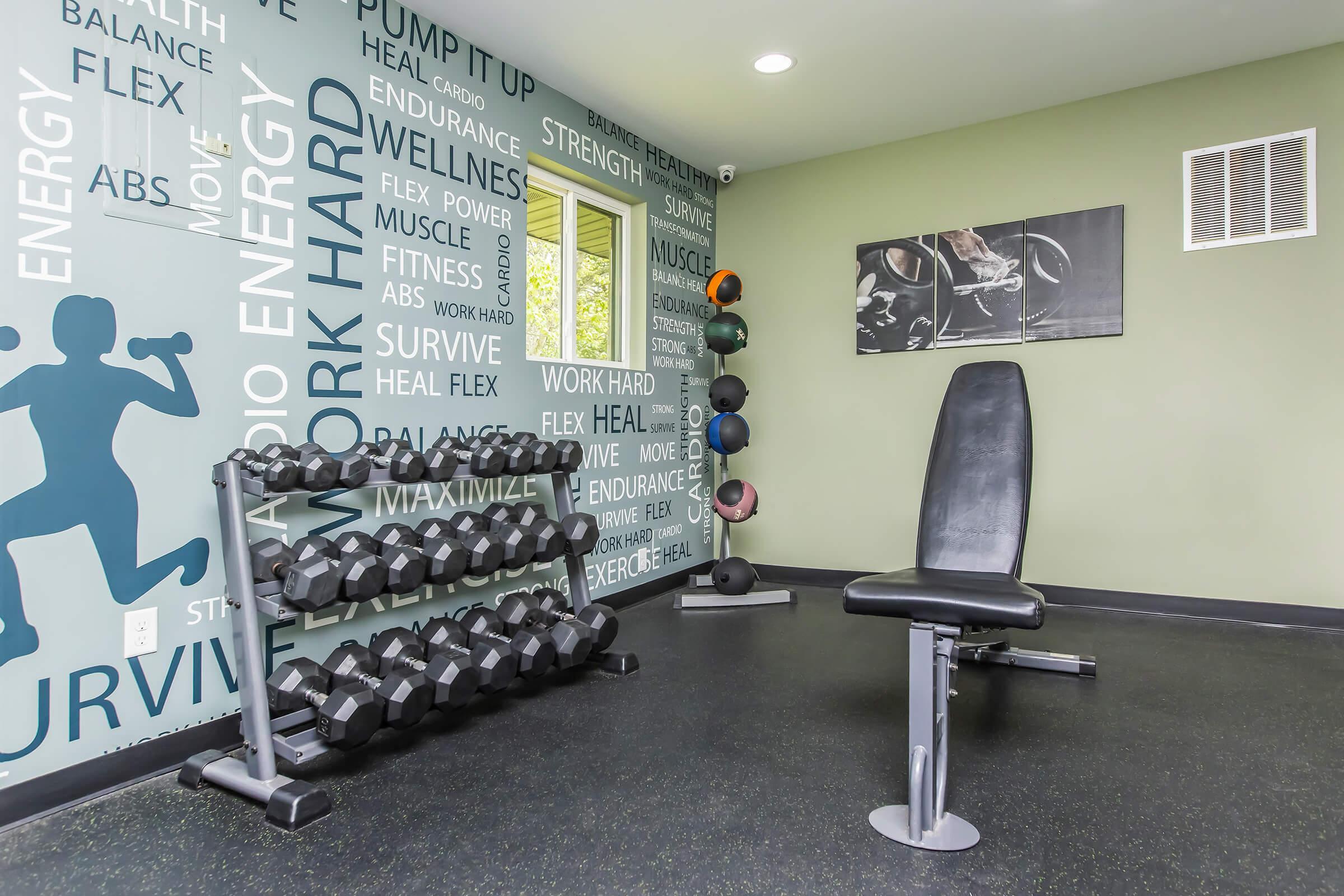 A well-equipped home gym featuring a dumbbell rack with various weights, a workout bench, and motivational wall decor. Large windows provide natural light, enhancing the uplifting atmosphere for fitness activities.