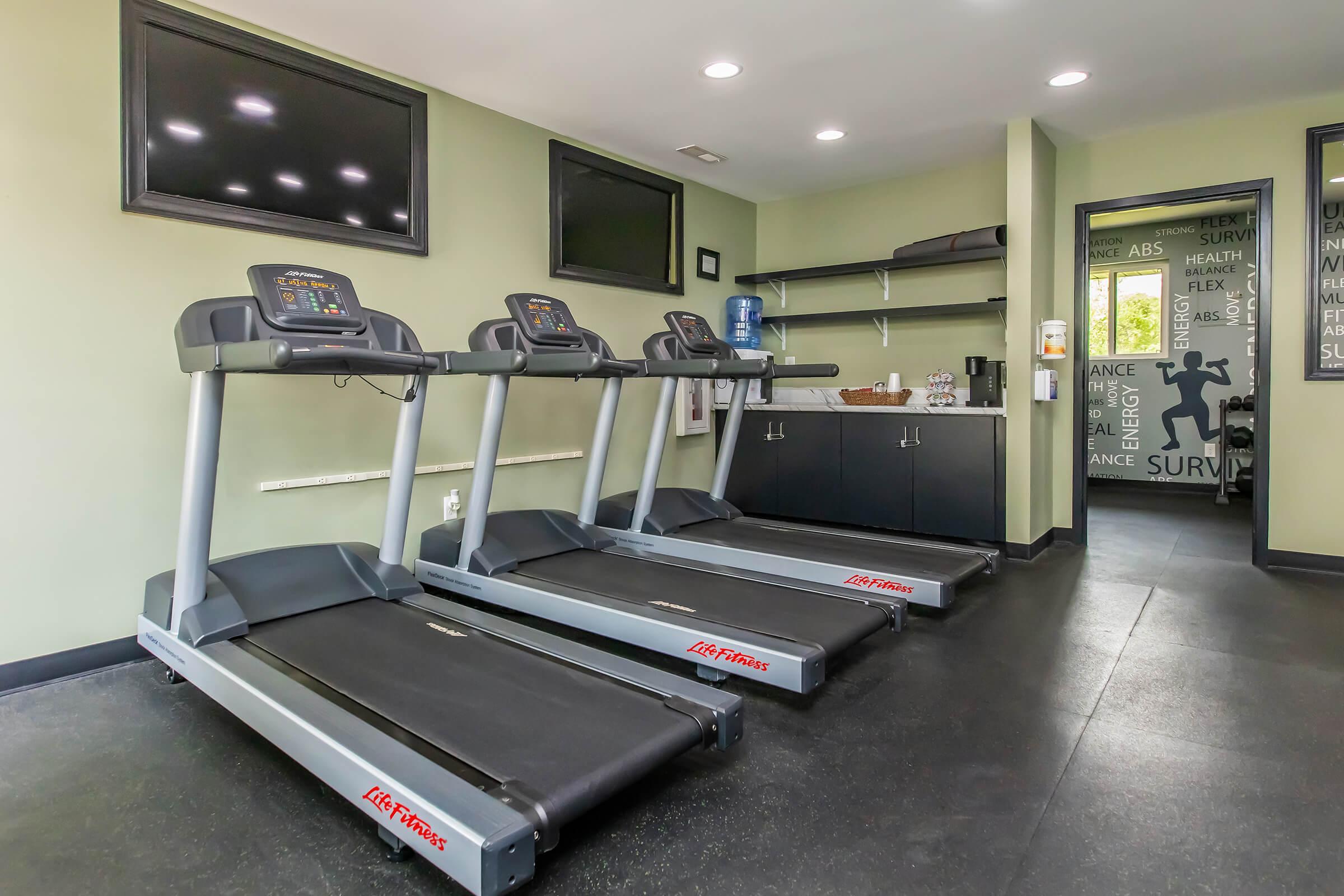 Three treadmills lined up in a modern gym space. The room features green walls, bright overhead lighting, and a small refreshment area with a water cooler and shelving. Two large screens are mounted on the wall, and there's a motivational mural in the background.