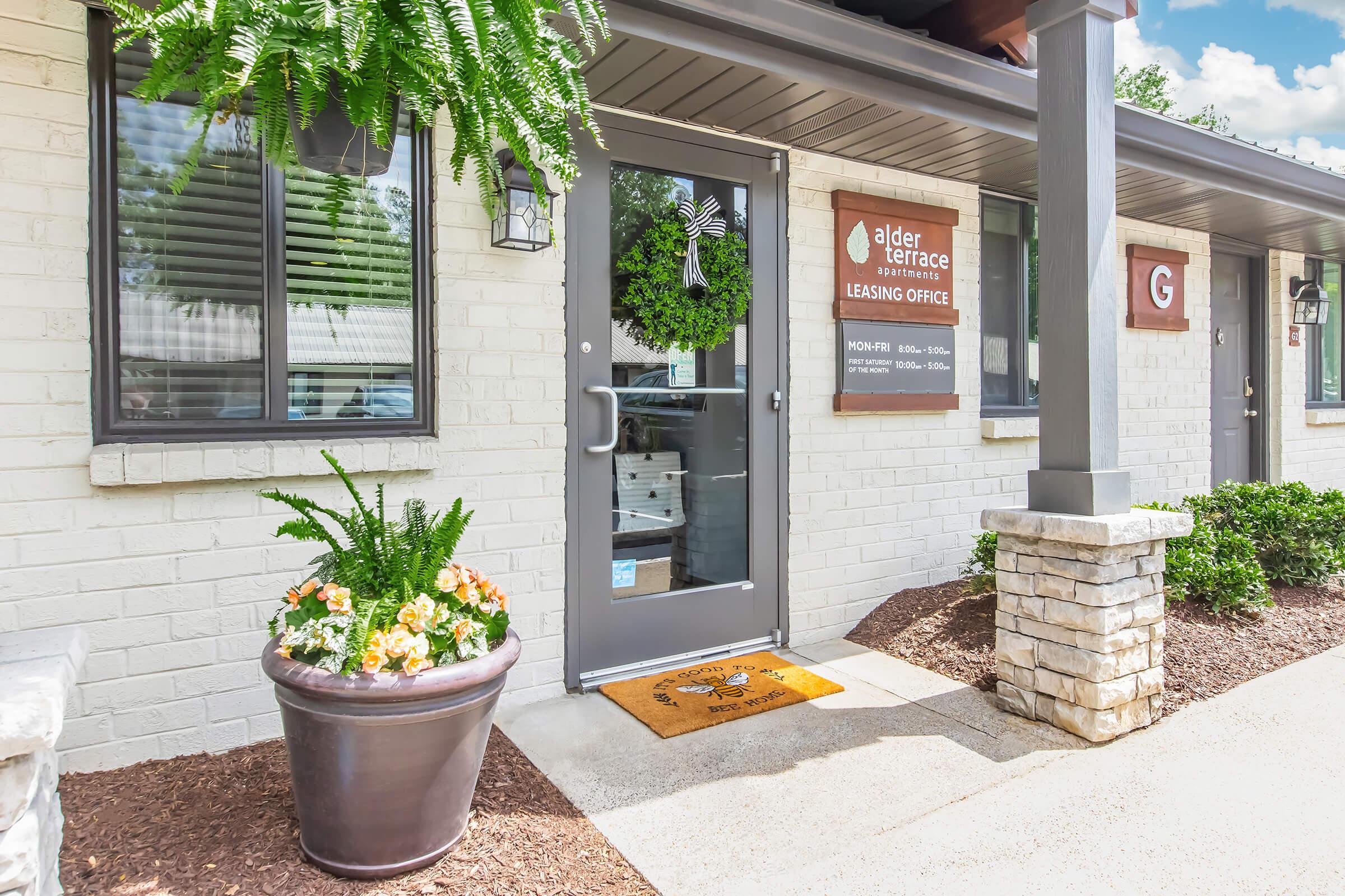A welcoming leasing office entrance with a dark gray door, adorned with a green wreath and a decorative welcome mat. Flanking the entrance are potted plants and a stone column, with a sign detailing office hours and leasing information visible beside the door.