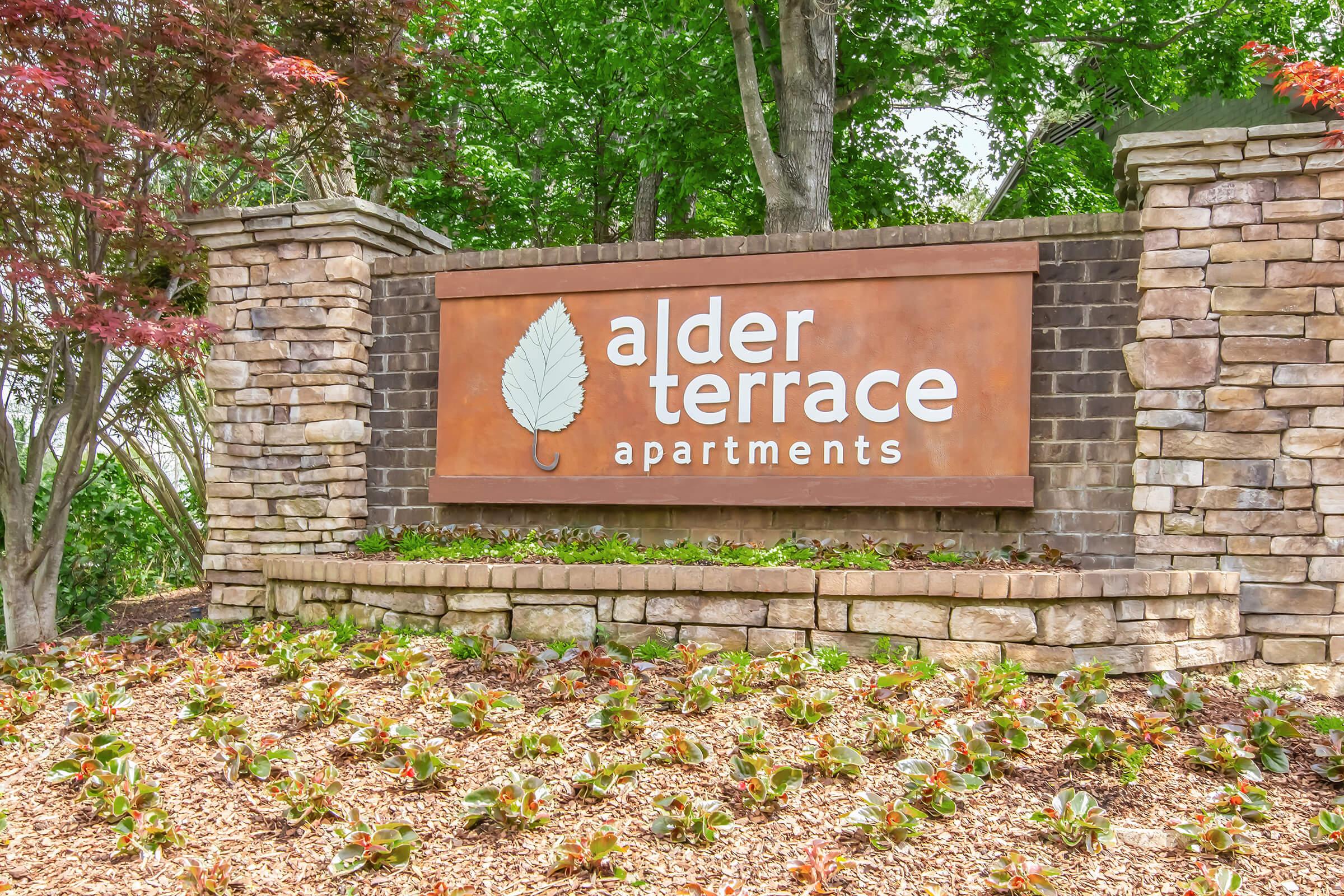 Sign for Alder Terrace Apartments featuring the name prominently displayed in white text on an orange background, surrounded by a stone wall. The area is landscaped with green plants and trees, creating a welcoming atmosphere.
