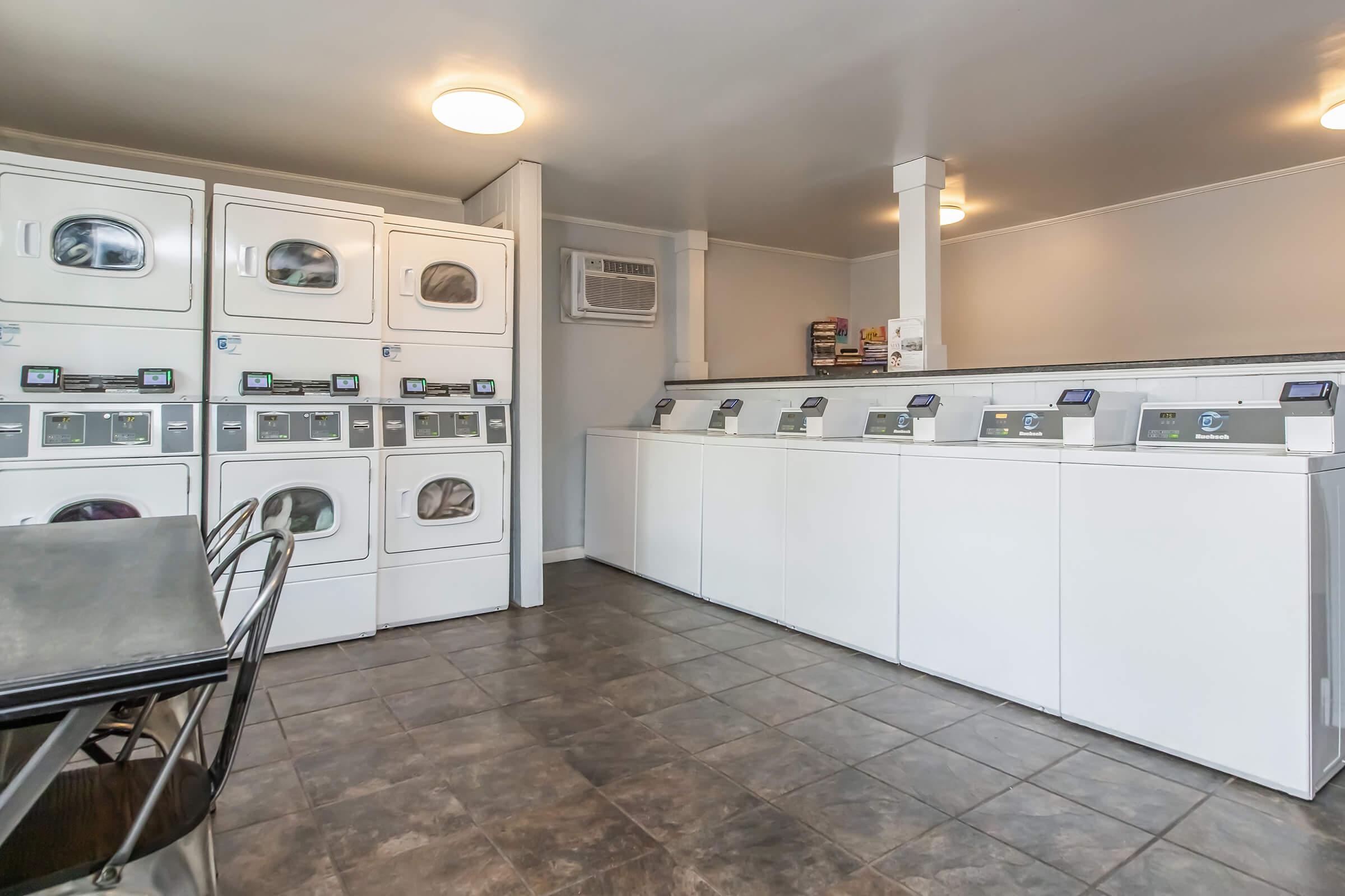 A clean laundry room featuring multiple stacked washing machines and dryers, along with folding tables. The space is well-lit with overhead lights and has tiled flooring. There is an air conditioning unit on the wall and a few laundry supplies on the counters.