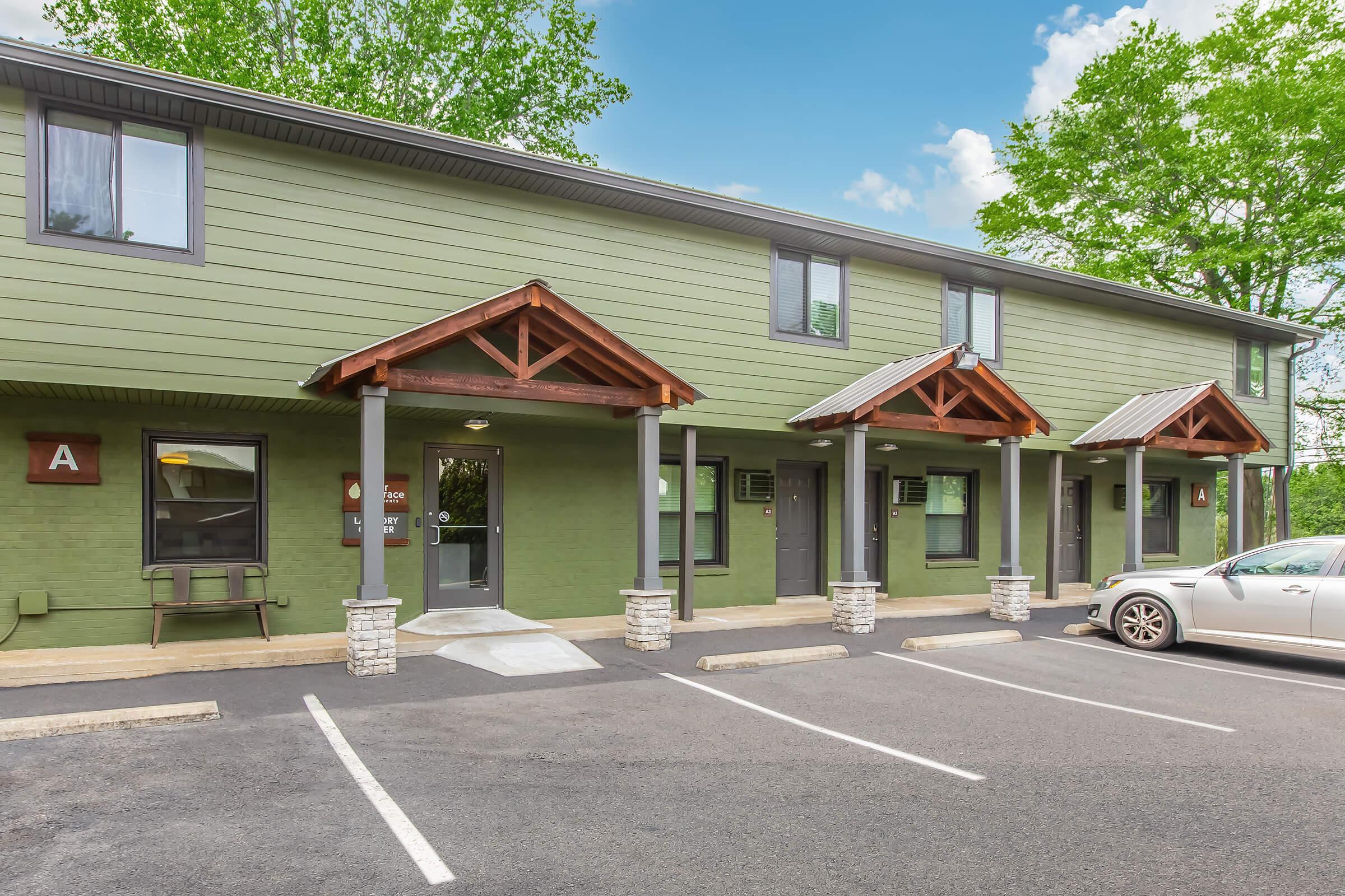 Exterior view of a two-story green building with wooden awnings over the entrance doors. The building features multiple units, a small bench, and a car parked in front. There are parking spaces and trees in the background, with a bright blue sky and clouds visible.