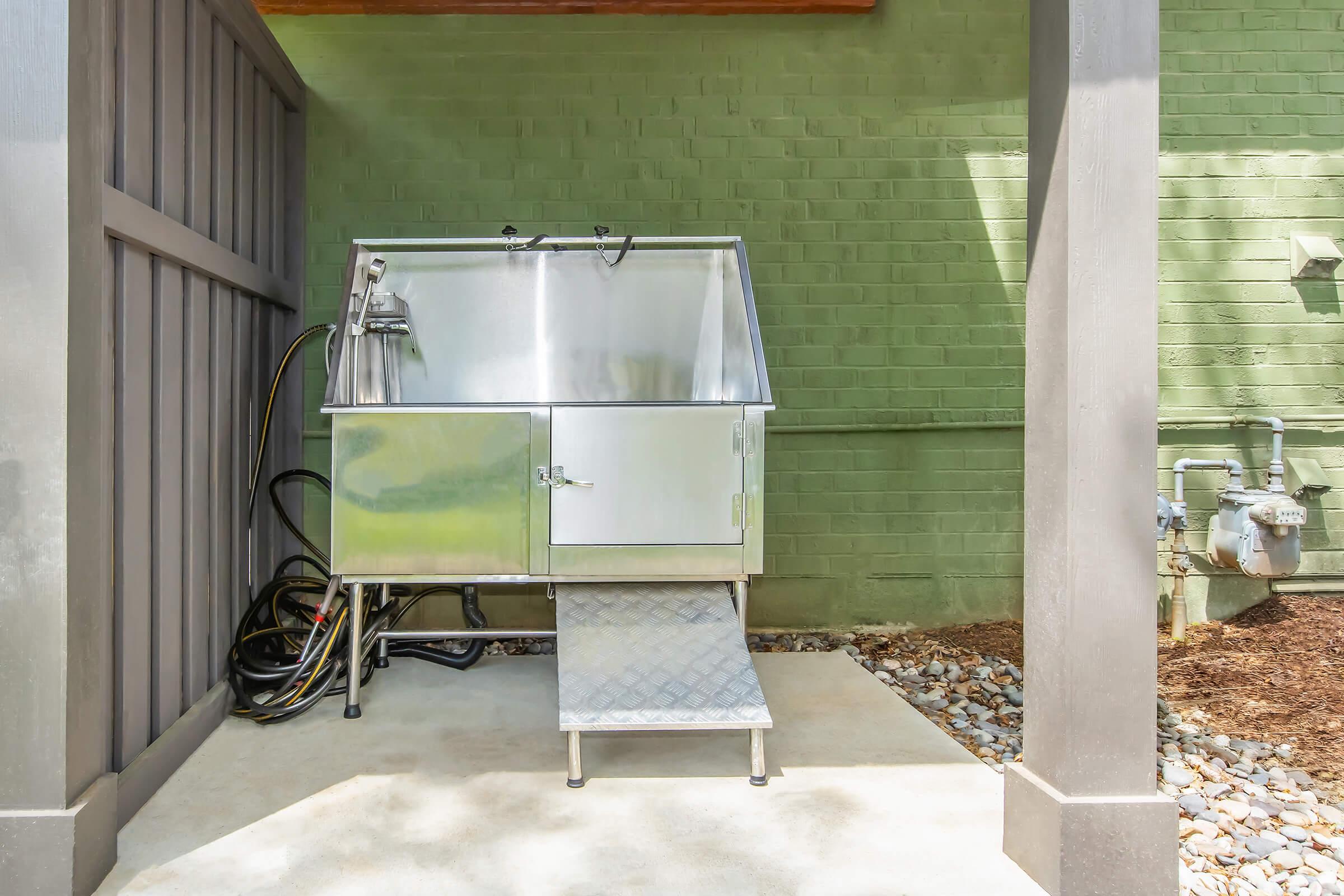 A stainless steel outdoor washing station with a sloped ramp, situated against a green brick wall. The station features a faucet and a closed storage compartment, with hoses connected nearby. The area is surrounded by a wooden structure and has a gravel surface.