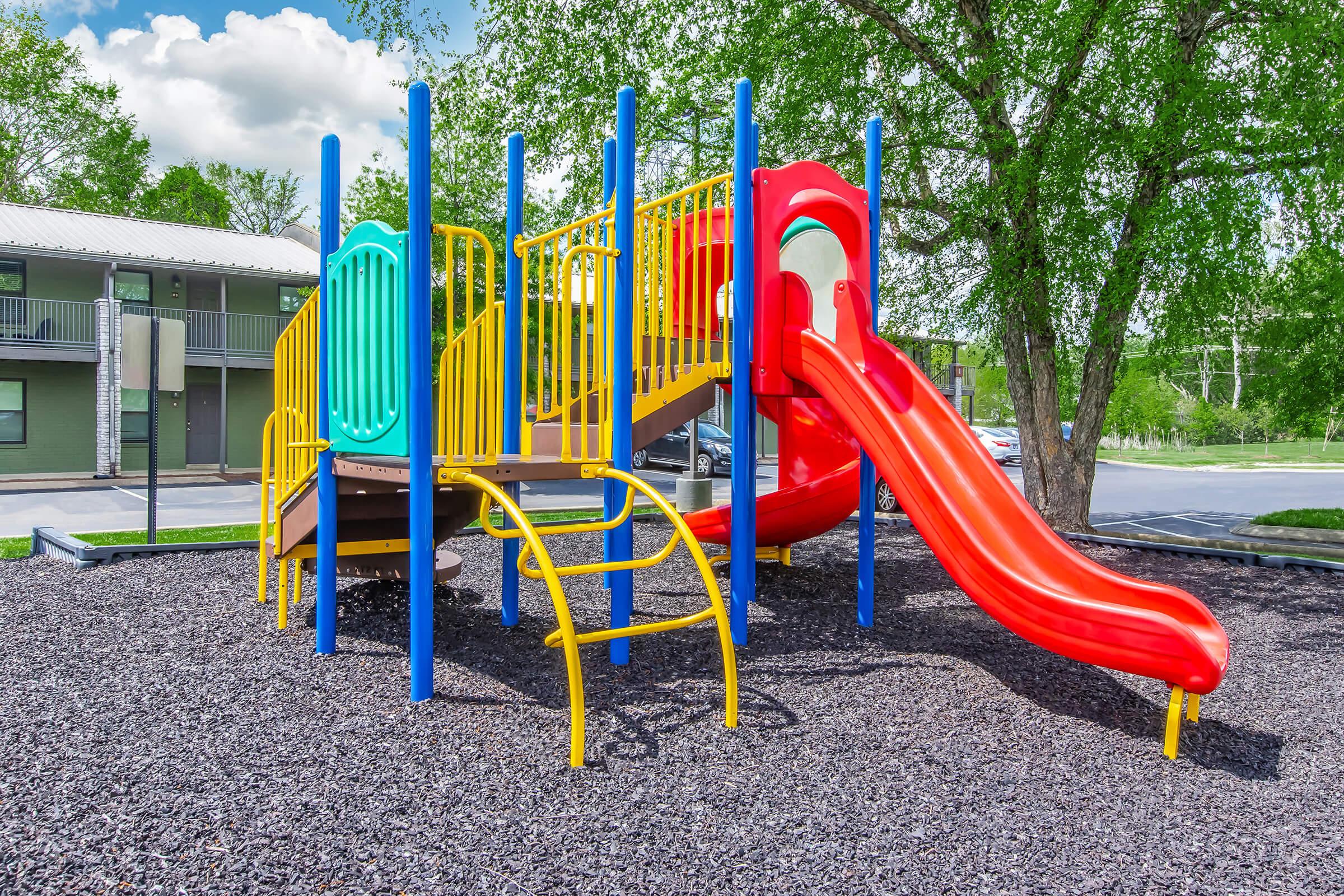 A colorful playground structure featuring a slide, climbing frame, and safety barriers set on a bed of dark mulch. The slide is bright red, while the frame includes yellow and blue elements. Surrounding the playground are trees and a parking area, with a building visible in the background. Bright blue sky with clouds overhead.