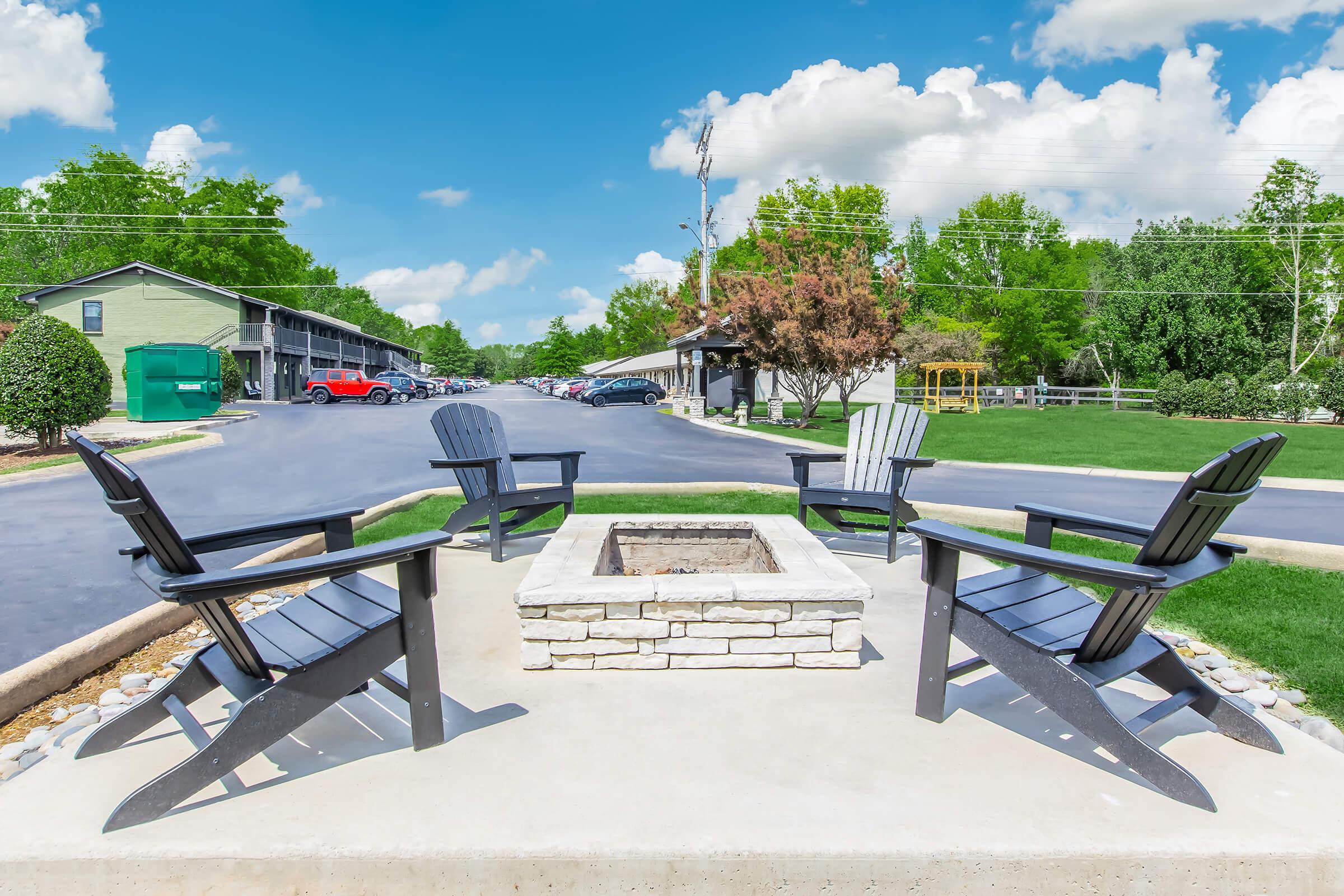 A cozy outdoor seating area featuring four black Adirondack chairs arranged around a stone fire pit. Surrounding greenery and a clear blue sky with fluffy white clouds create a welcoming atmosphere. In the background, there are residential buildings and a parking lot.