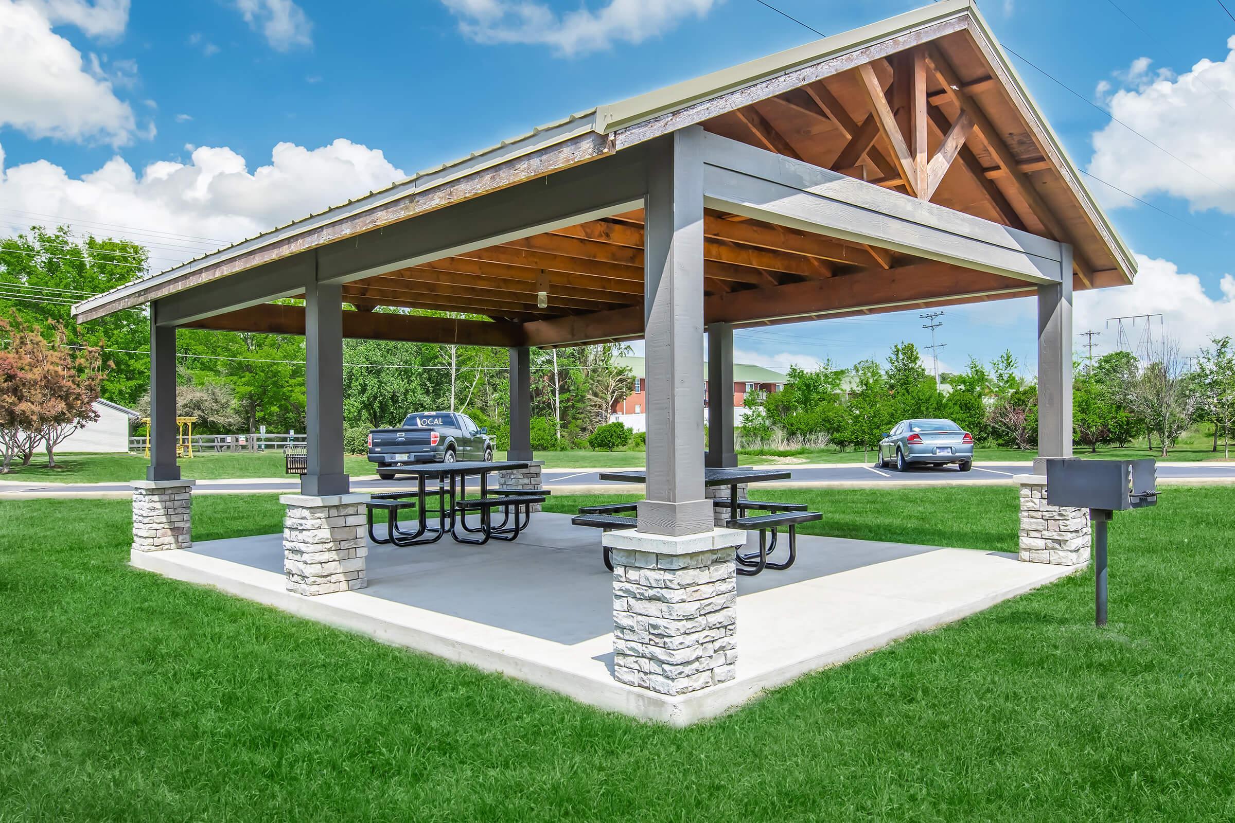 A well-lit park pavilion with a wooden roof and stone columns, featuring several picnic tables underneath. Lush green grass surrounds the pavilion, and there are parked cars and trees in the background under a partly cloudy sky.