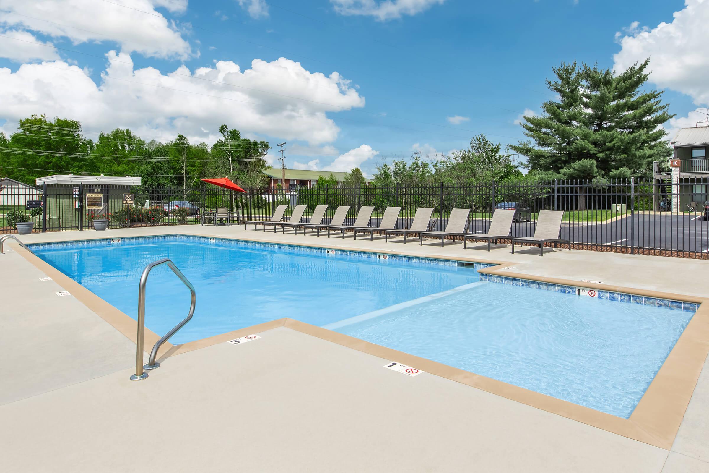 A bright outdoor pool area featuring a large swimming pool and a shallow wading section. Surrounding the pool are multiple lounge chairs lined up neatly, with a few shaded by red umbrellas. Lush green trees and landscaping are visible in the background under a partly cloudy blue sky.