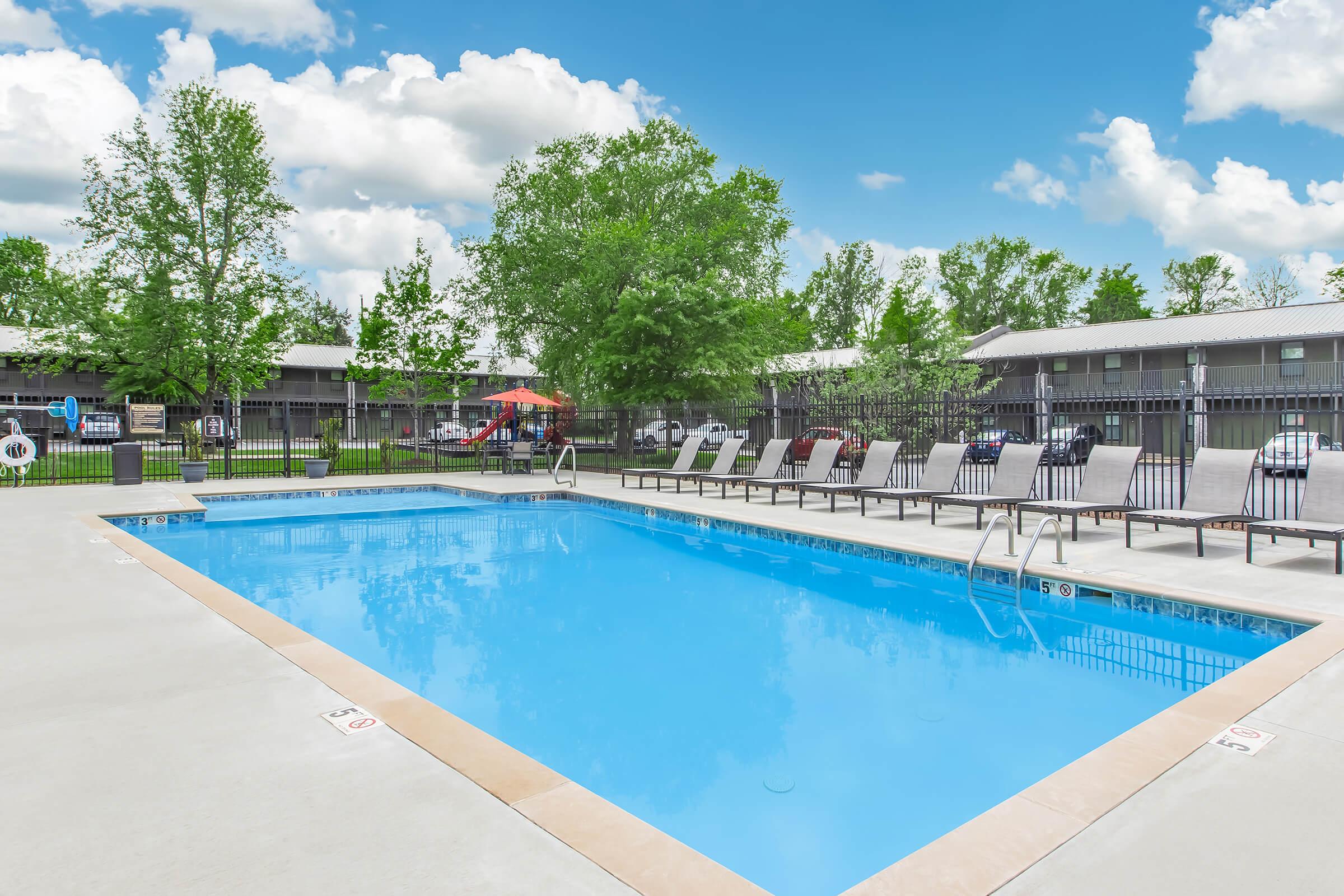A sparkling blue swimming pool surrounded by lounge chairs and greenery. The pool area features a fenced perimeter, with shaded seating under umbrellas. Light clouds drift across a bright blue sky, and nearby buildings provide a residential backdrop.