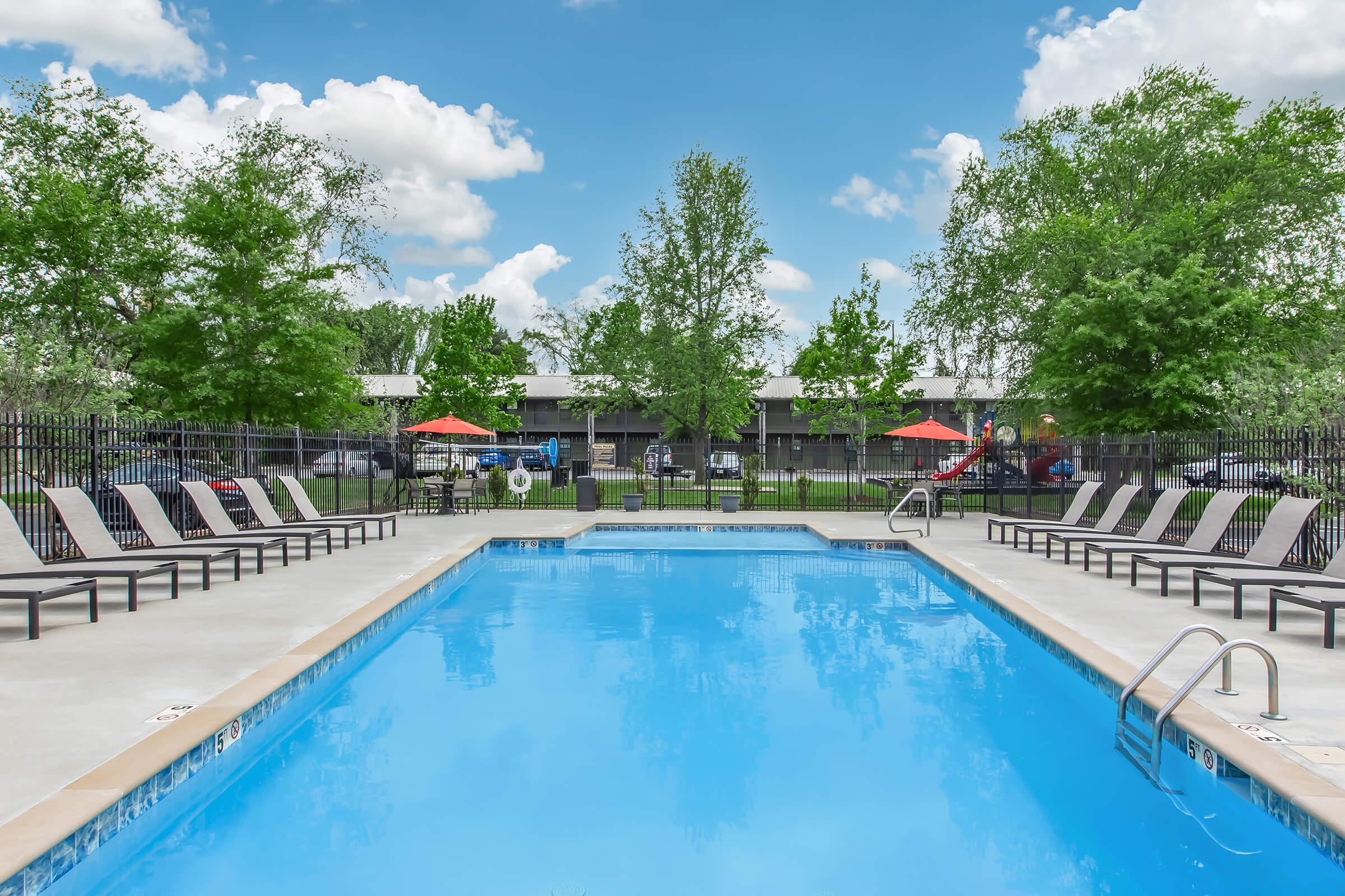 A clear swimming pool surrounded by lounge chairs, with trees and a blue sky in the background. Red umbrellas provide shade, and there appears to be a playground area in the distance. The scene conveys a relaxing outdoor atmosphere.