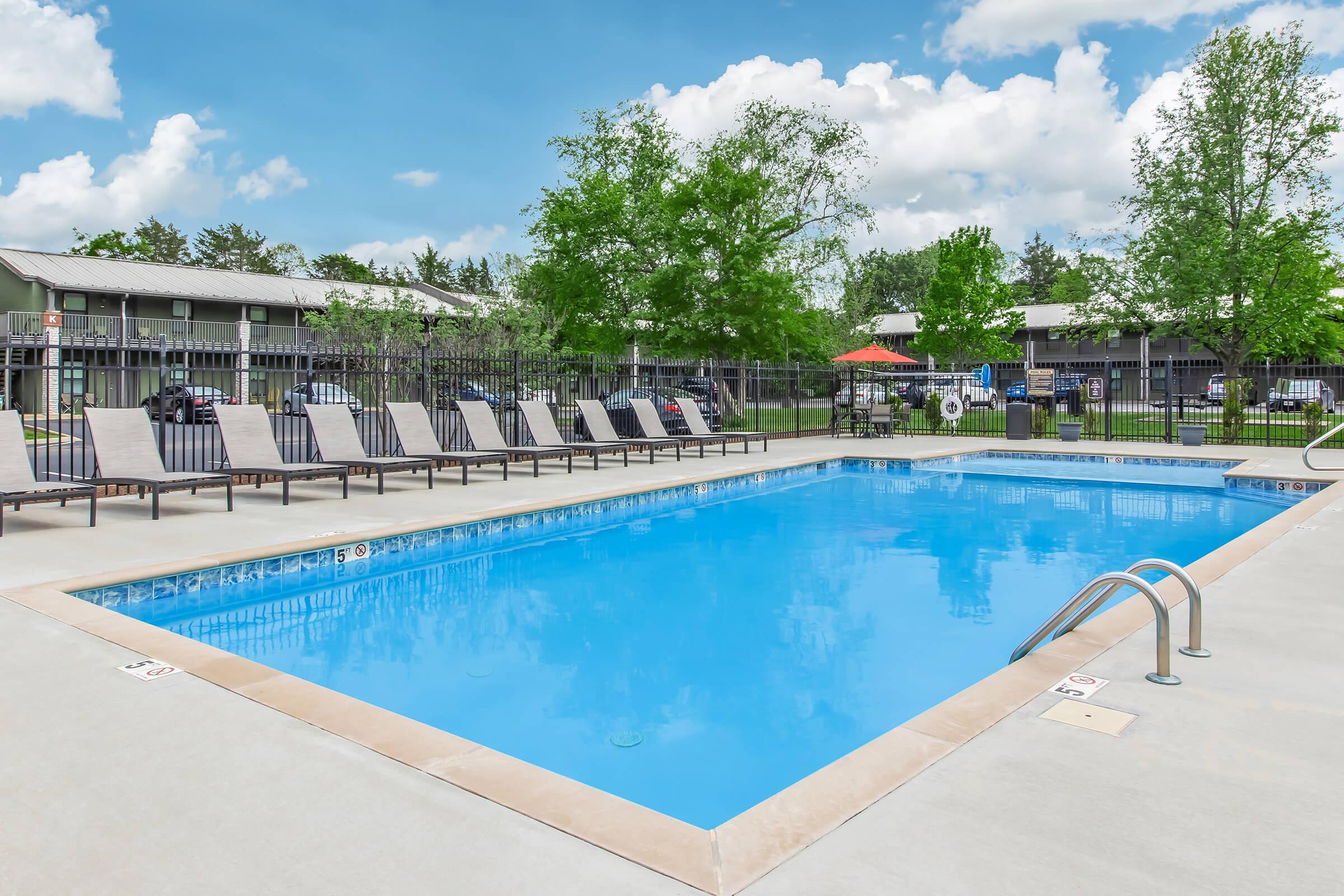 A clear blue swimming pool surrounded by lounge chairs under a bright sky with fluffy clouds. In the background, there are trees and a building structure. The pool area is well-maintained, and there is a safety ladder on one side.