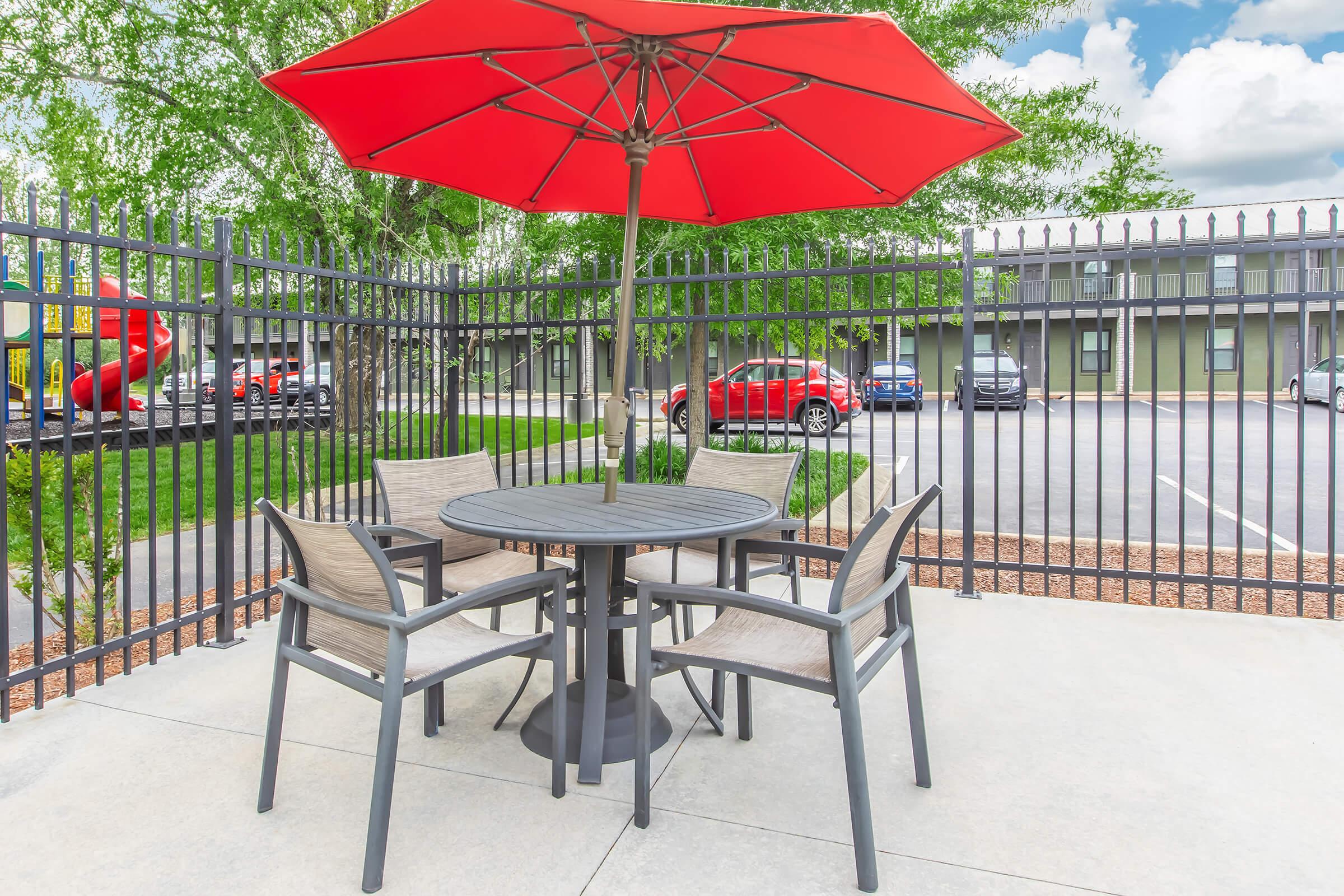 A patio setting featuring a round table with four chairs under a bright red umbrella, surrounded by a black fence. In the background, there are several cars parked and greenery visible, creating a relaxed outdoor atmosphere.