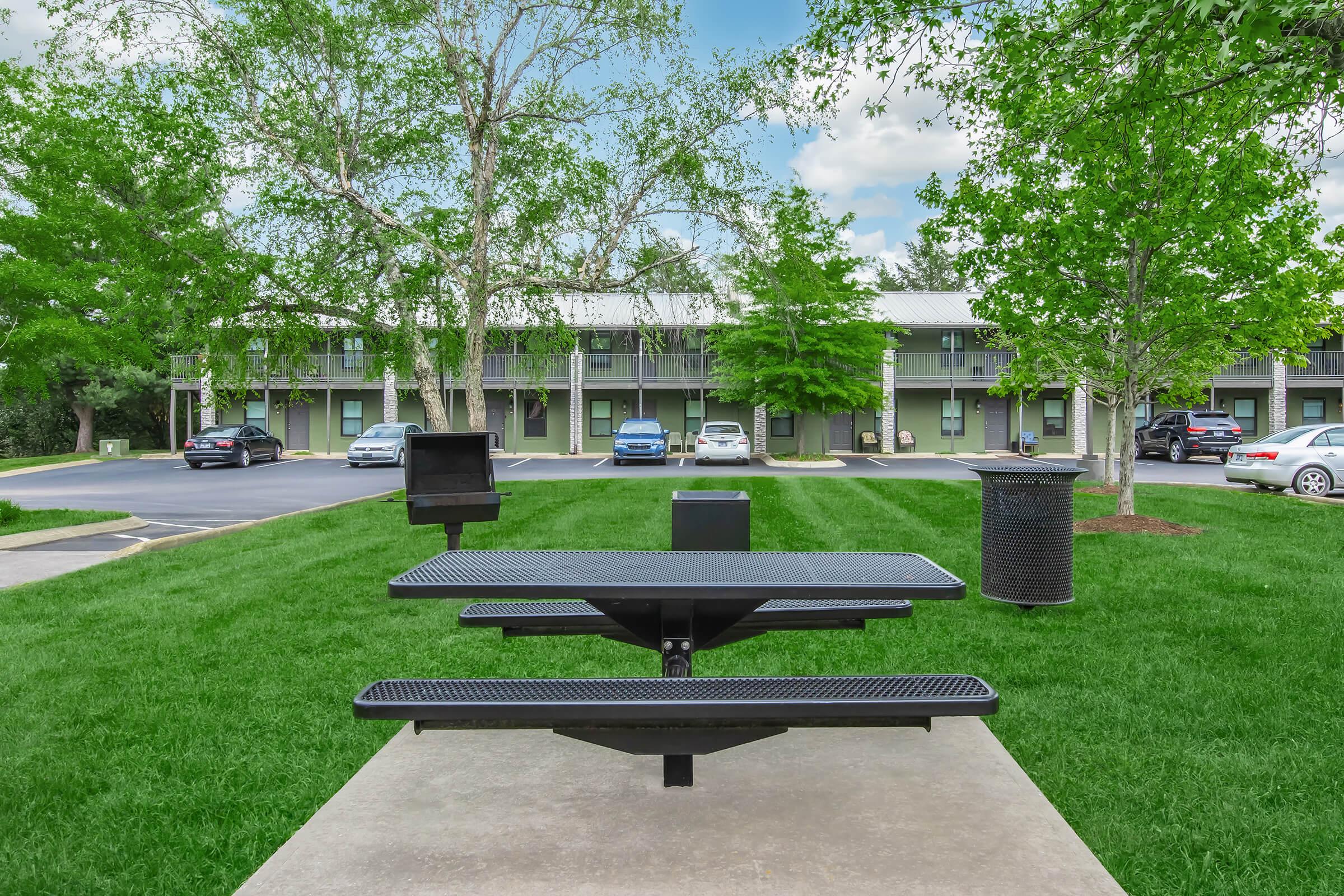 A picnic area featuring a black metal table and grill, set on green grass. In the background, there are multiple trees and a hotel with parked cars. The scene is bright and well-maintained, showcasing a relaxing outdoor space.