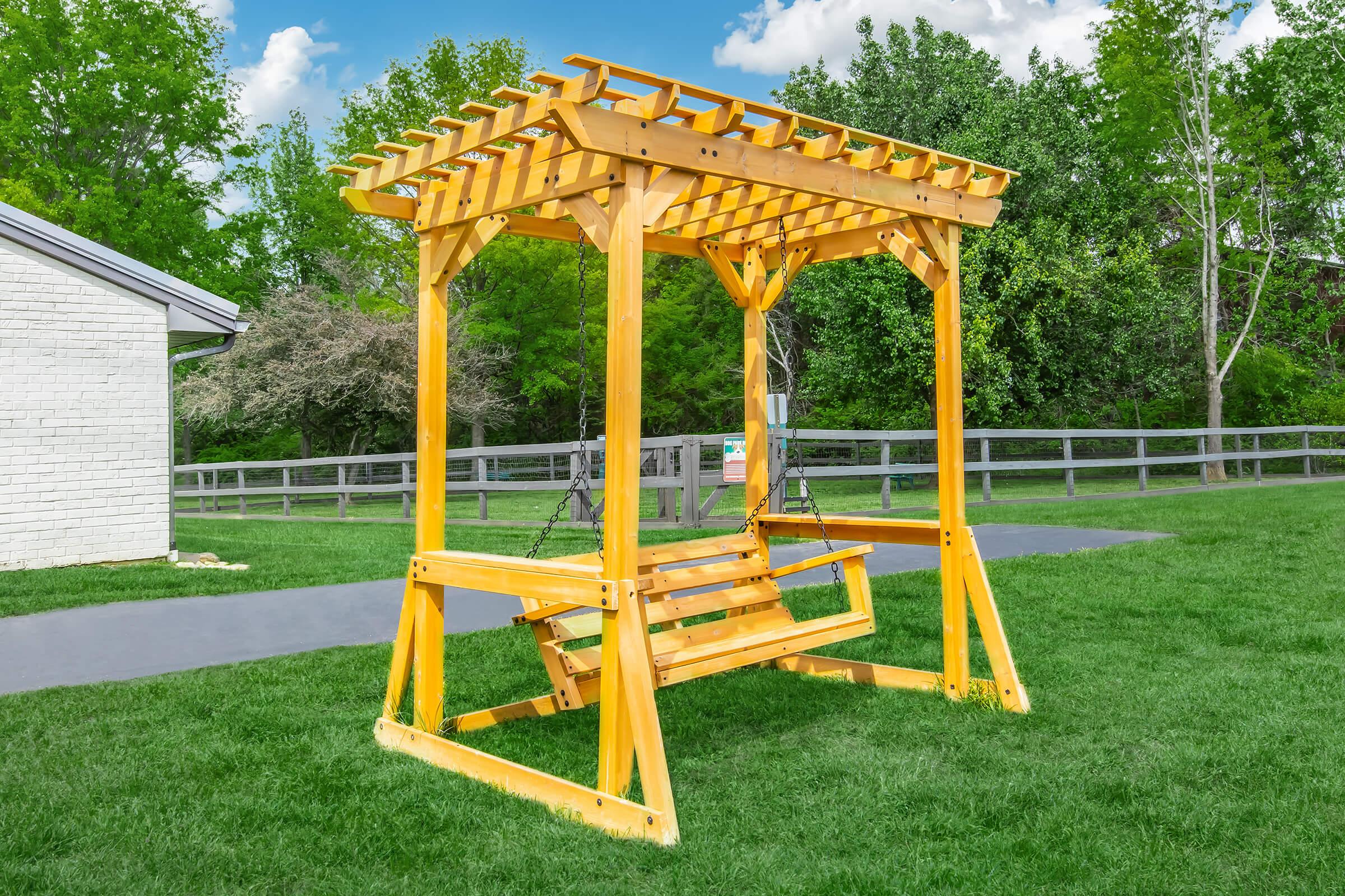 A wooden swing set with a pergola-style roof, placed on a grassy lawn. It features two swings suspended from the structure, surrounded by trees and blue skies, creating a serene outdoor environment.