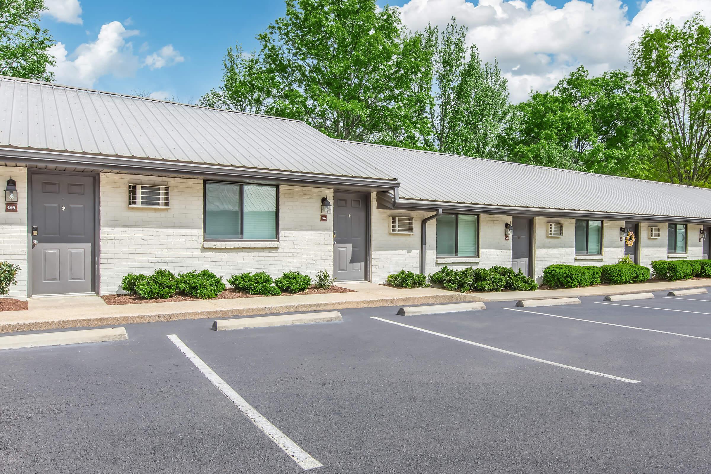 A row of motel rooms with gray doors and white brick exteriors. The building features a metal roof and surrounded by well-maintained landscaping, including small bushes. A parking lot with empty spaces is visible in front, set against a backdrop of green trees and a blue sky with scattered clouds.