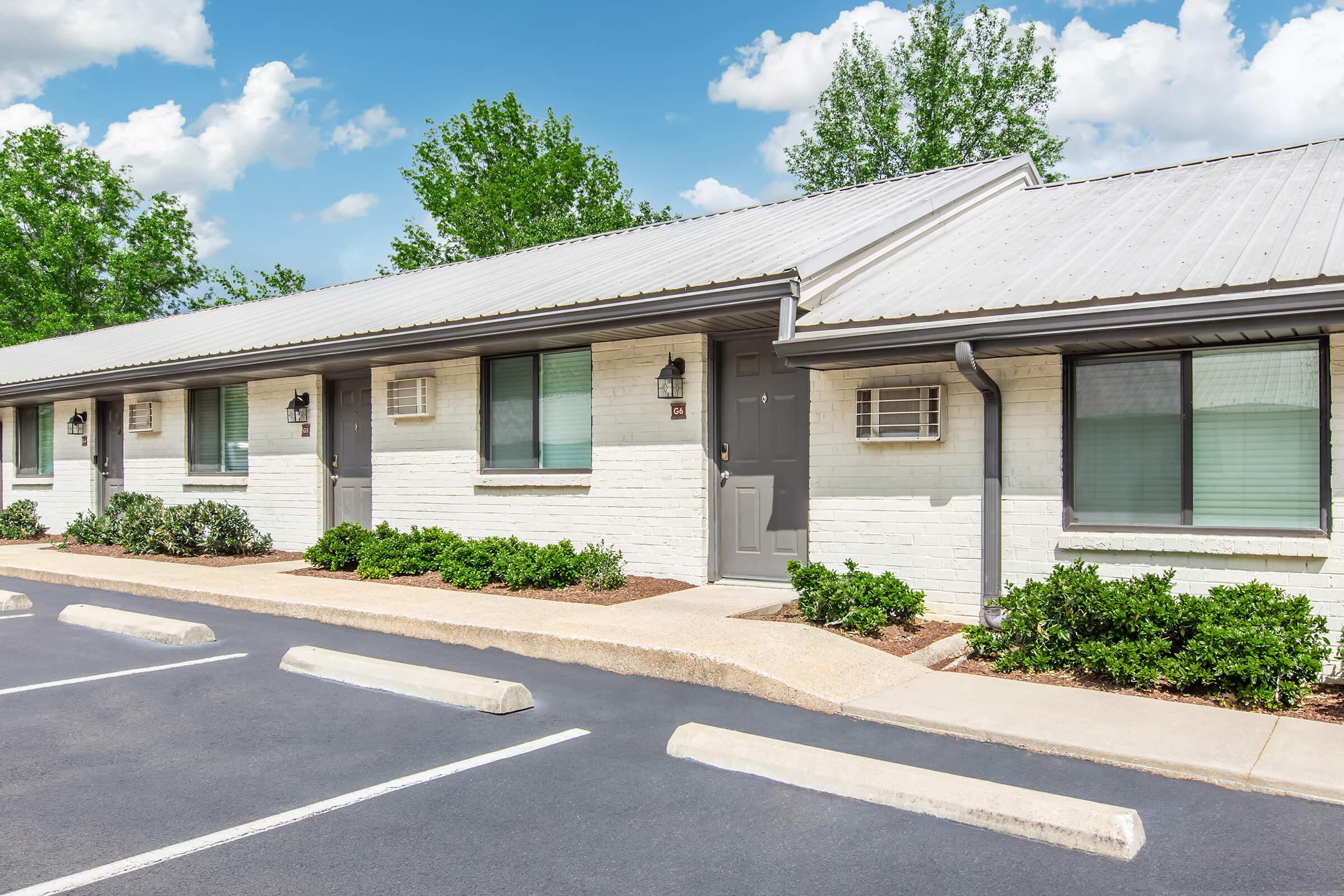 A row of single-story motel rooms with gray doors and windows, surrounded by green shrubs. A paved parking area is visible in front, with several empty parking spaces. The background features trees and a clear blue sky with scattered clouds.