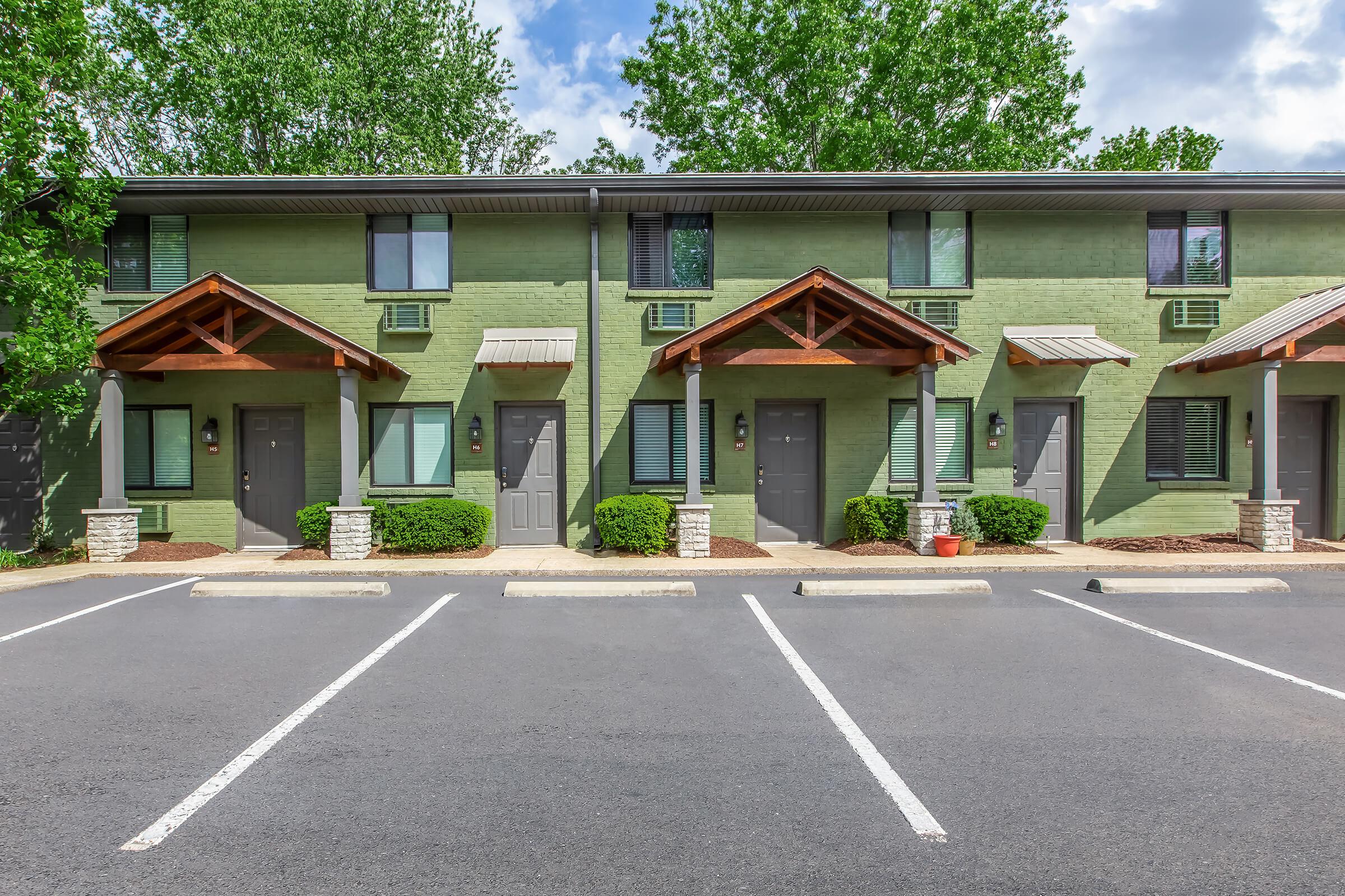 A row of green townhomes with wooden awnings and stone accents. The entrance of each unit features a door and windows, surrounded by neatly trimmed bushes. A parking area with marked spaces is visible in front, under a partly cloudy sky with trees in the background.