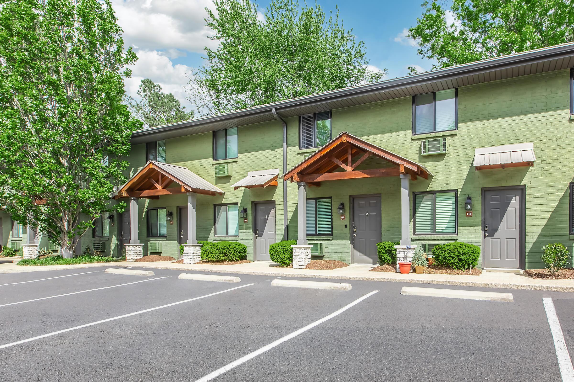 A row of green-painted townhouses with wooden awnings, situated in a well-maintained area. Trees and shrubs surround the buildings, and a parking lot with marked spaces is in the foreground. The sky is partly cloudy, adding a bright atmosphere to the scene.