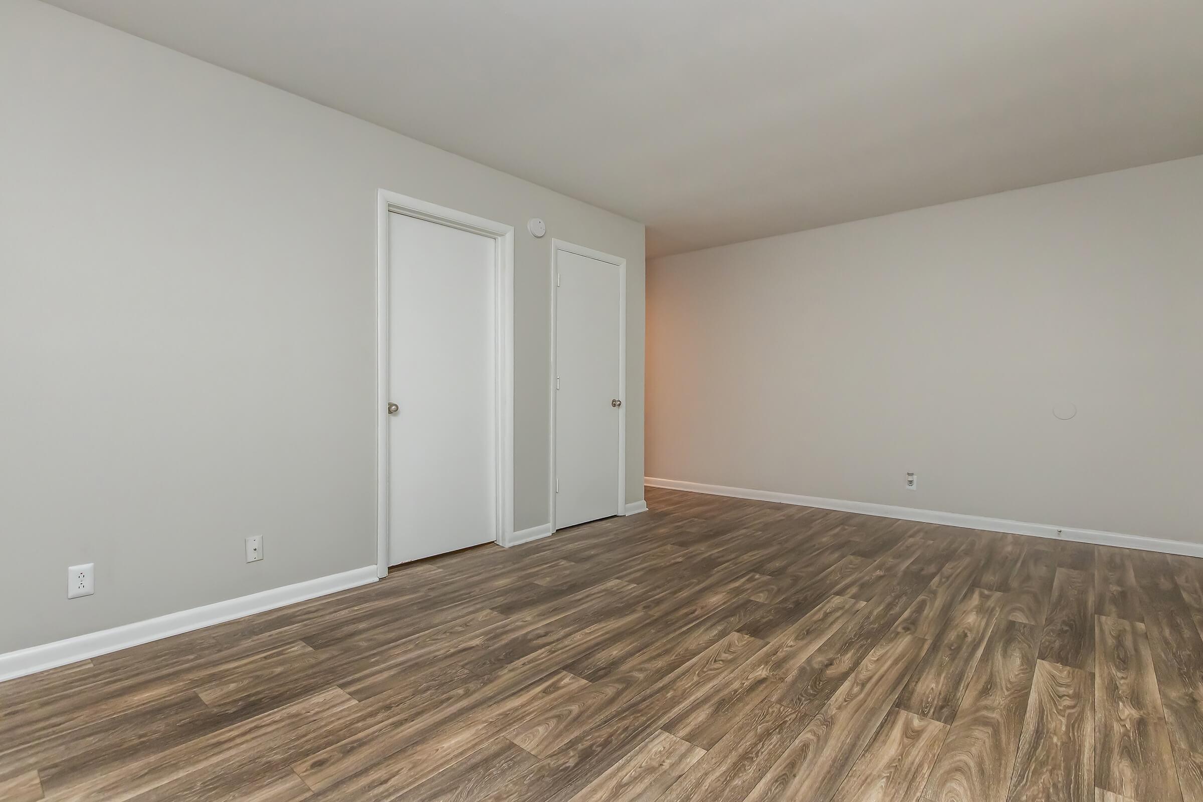 A spacious, empty room with light-colored walls and new wood laminate flooring. Two closed white doors are visible on the left side of the image. The room is well-lit, emphasizing its clean, neutral aesthetic, and there's no furniture present.