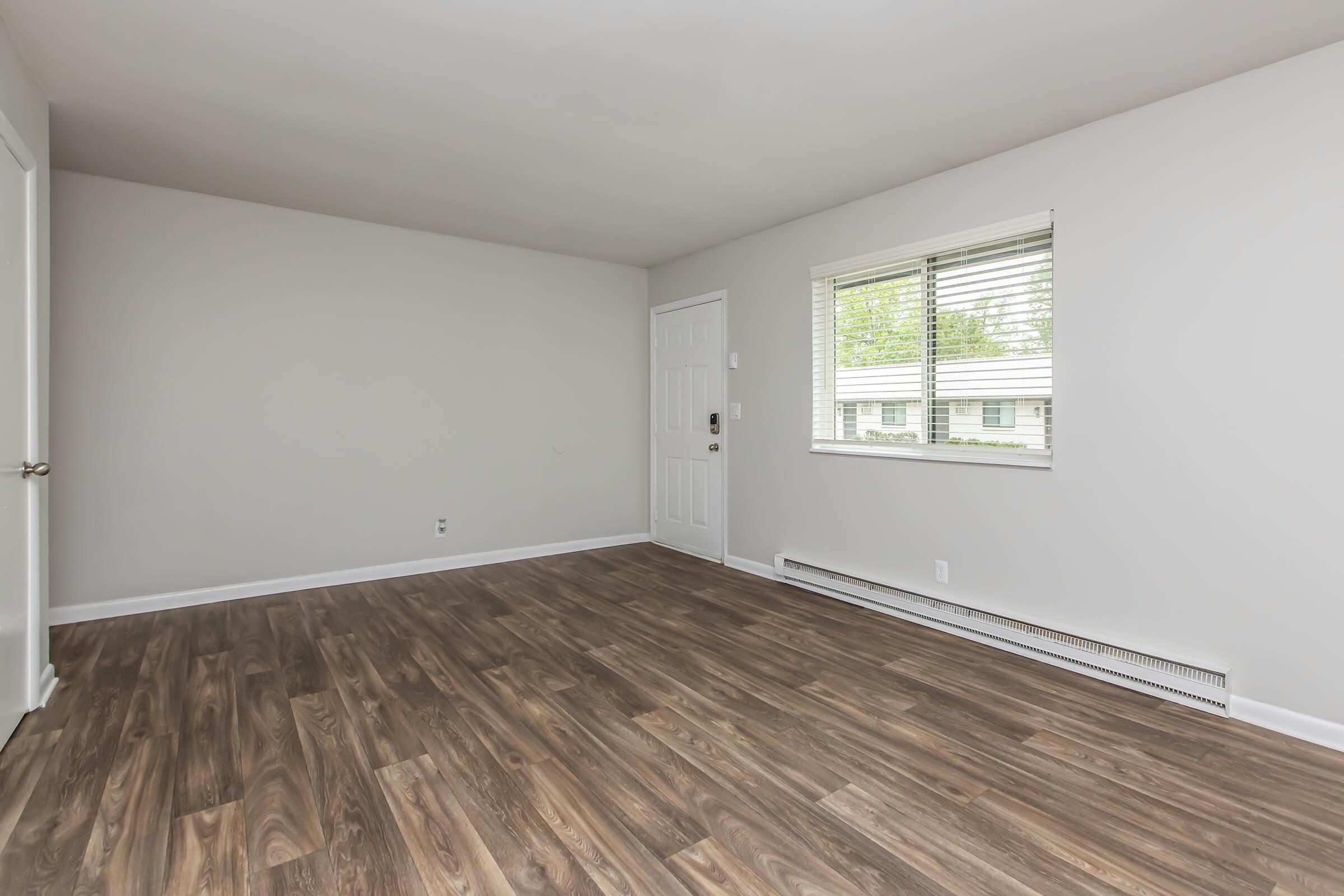 A minimalistic interior view of a living room featuring light gray walls, a white door, and a large window with blinds. The floor is a warm-toned wood laminate, and the space is empty and well-lit, suggesting a fresh and spacious environment.