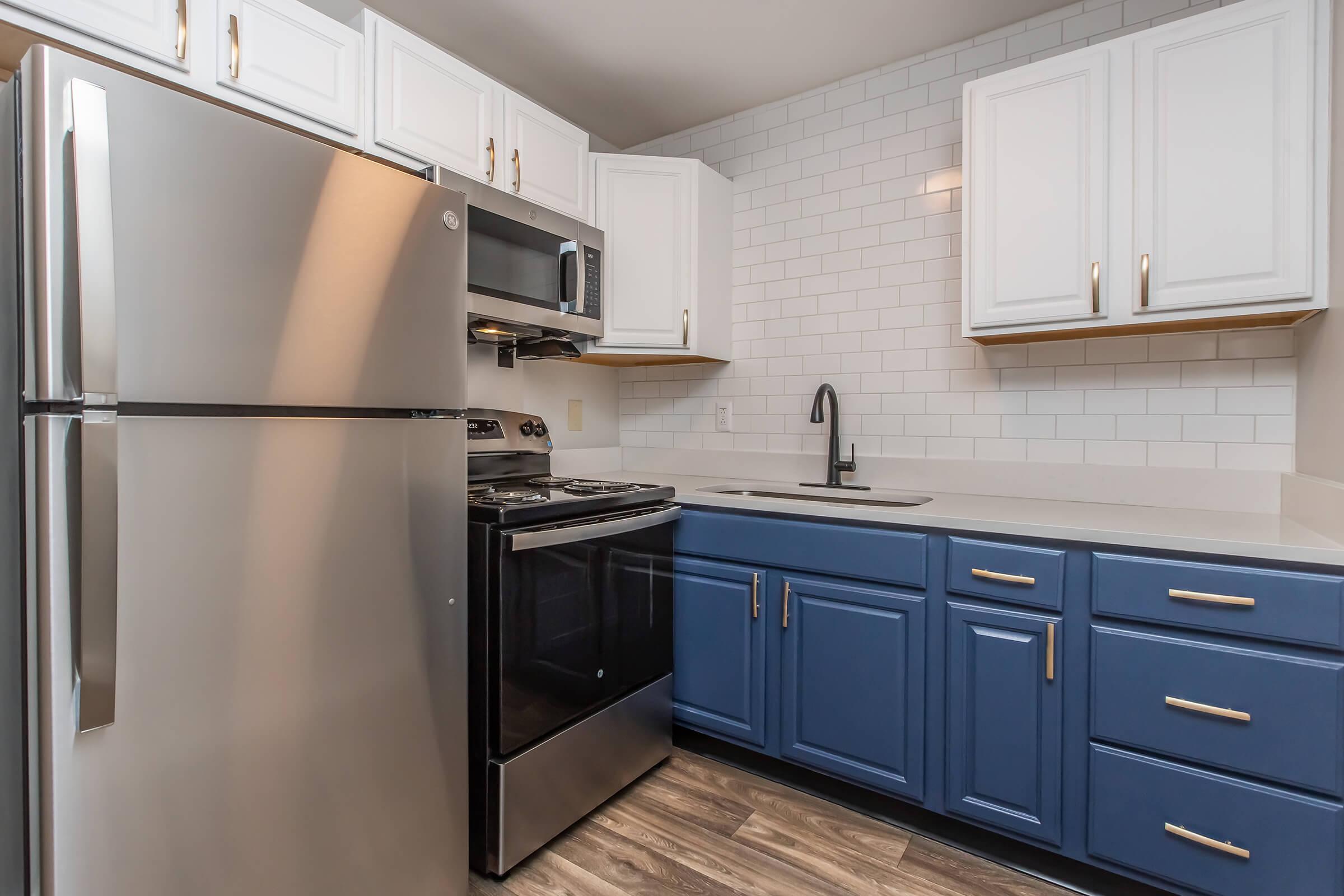 Modern kitchen featuring stainless steel appliances including a refrigerator and stove, white upper cabinets, blue lower cabinets, and a sleek countertop with a sink. The backsplash consists of white subway tiles, providing a clean and stylish look. The flooring is a light wood-like material.
