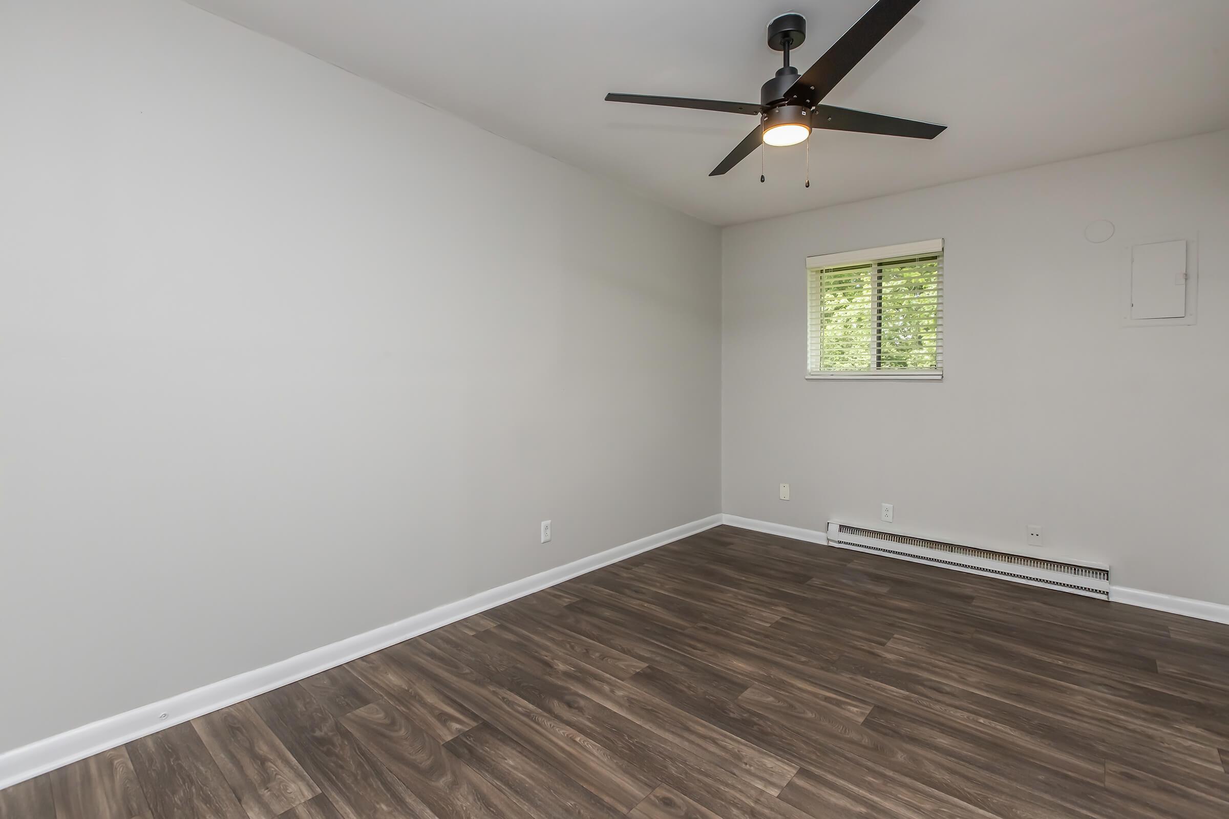 A spacious, empty room featuring light gray walls, a ceiling fan with a light fixture, and a window providing natural light. The flooring is made of dark wood laminate, and there is a heating unit along the wall. The overall atmosphere is clean and modern.