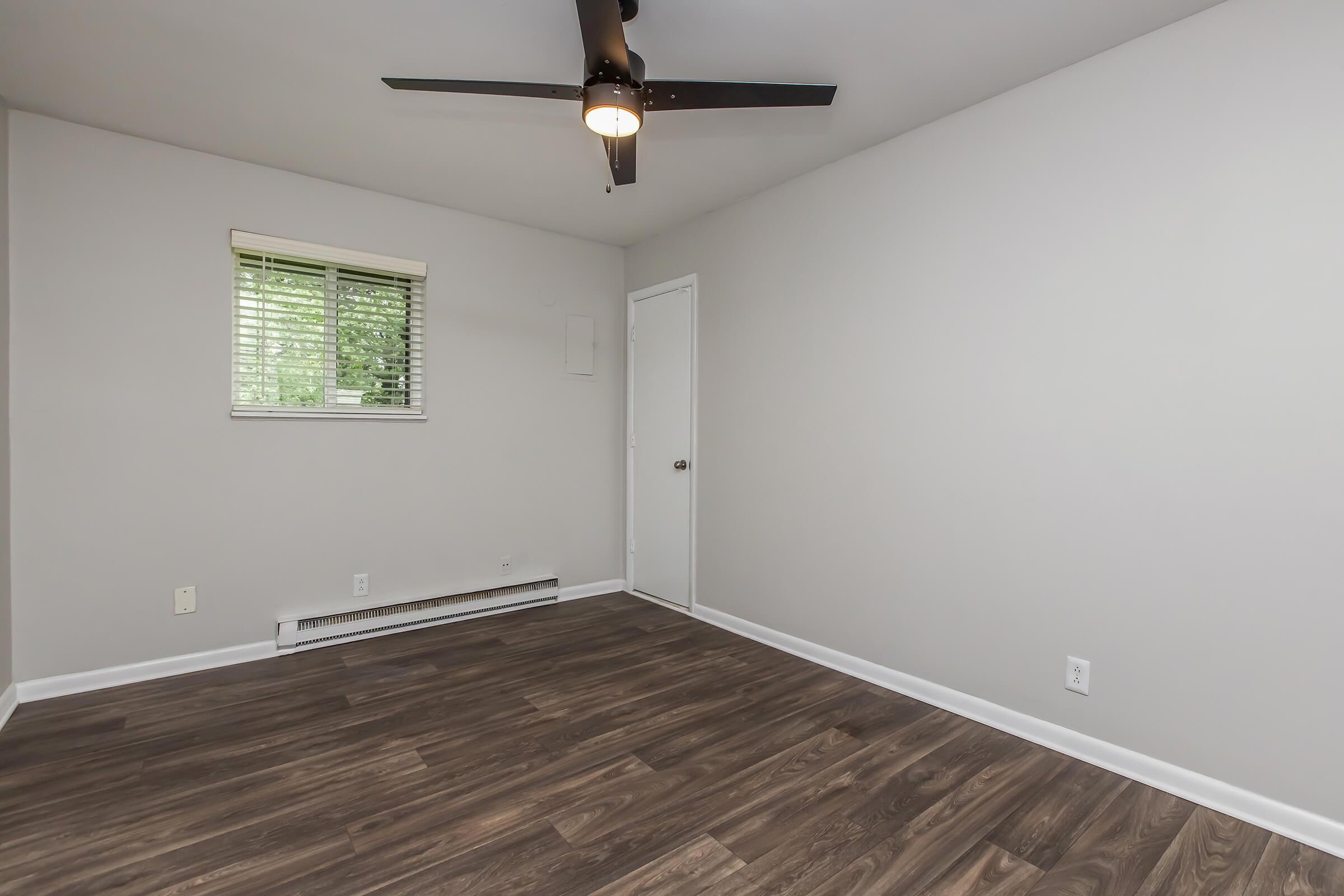 A spacious, empty room with light gray walls and dark hardwood flooring. A ceiling fan is mounted above, and there is a window with blinds allowing natural light. A closed door is visible on one wall, enhancing the room's minimalist design. The overall atmosphere is bright and airy.