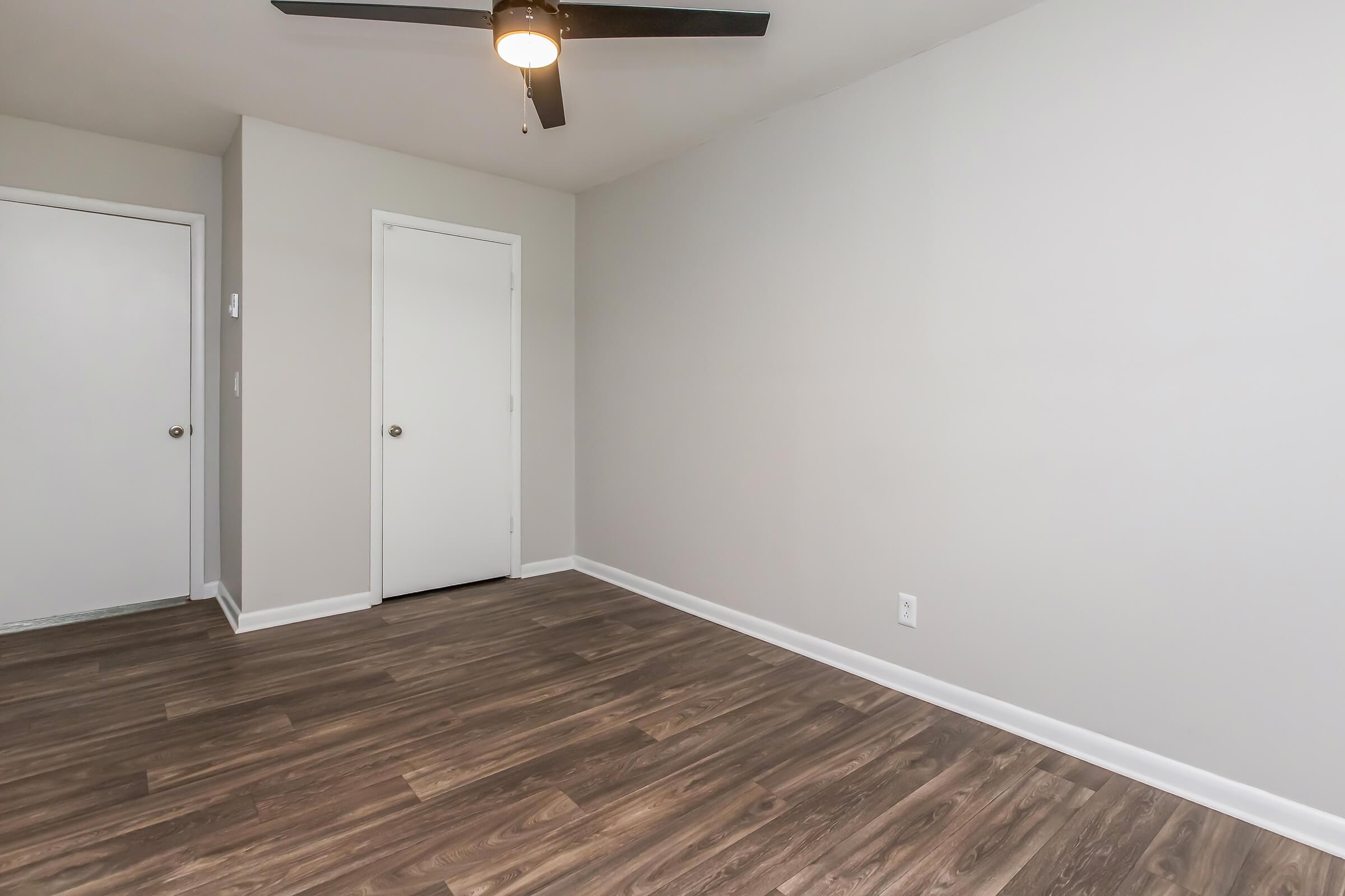 An empty room with a light-colored wall and dark wood flooring. A ceiling fan is mounted on the ceiling, and there are two closed doors: one leading to a closet and the other to another room. The space appears clean and well-lit, suggesting it is freshly renovated or maintained.
