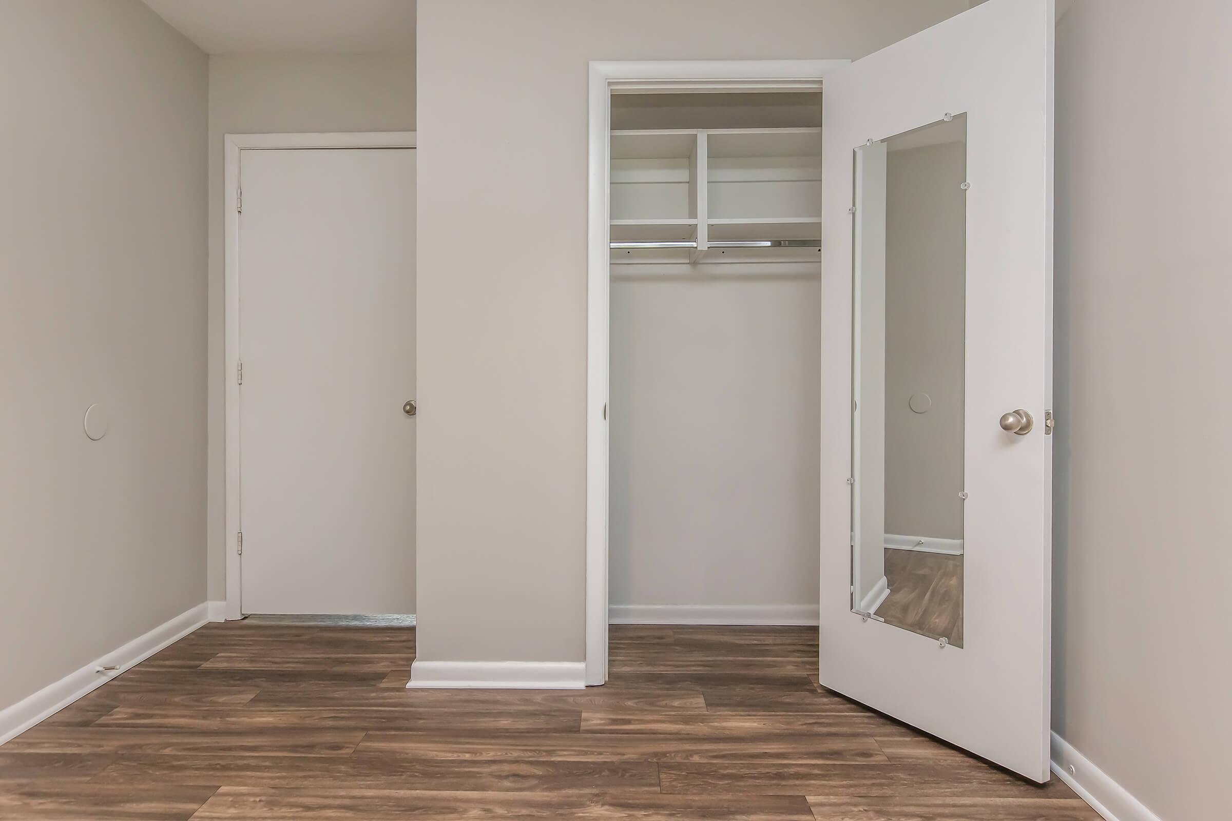 A well-lit, minimalist room featuring two doors—one leading to a closet with shelves and hanging space, and the other a standard door. The walls are painted light gray, and the floor is made of wood laminate, creating a clean and modern aesthetic.