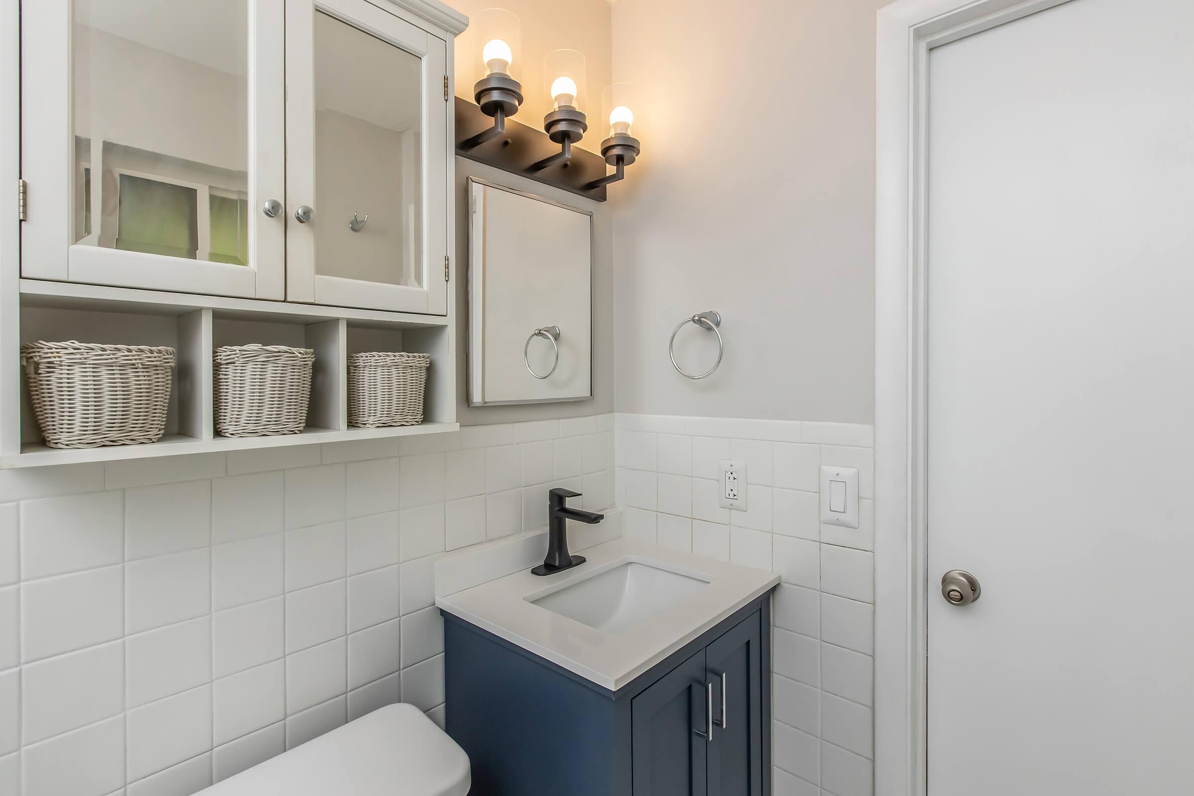 A modern bathroom featuring a wall-mounted mirror, a small sink with a dark blue cabinet beneath, and three woven baskets on a shelf. The wall is tiled in white, with a light fixture above the mirror providing soft illumination. A door can be seen leading out of the space.