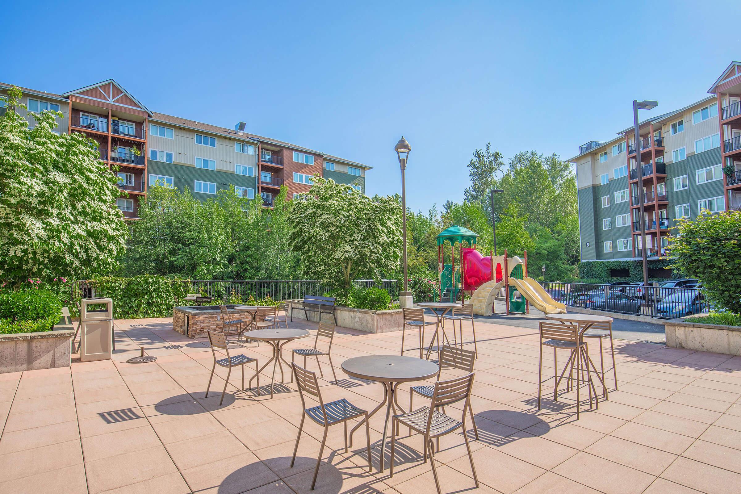 A sunny outdoor plaza featuring several metal tables and chairs, surrounded by greenery and trees. In the background, there are residential buildings with balconies. To the side, there's a playground with a slide and climbing structures, creating a vibrant, family-friendly atmosphere.