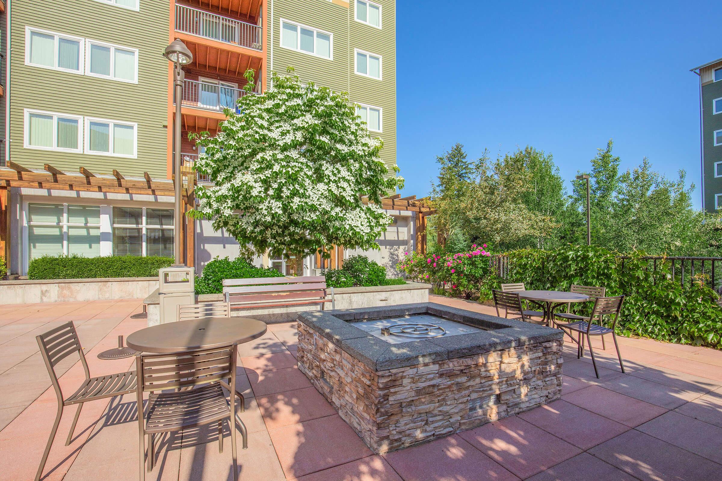 Outdoor seating area with metal tables and chairs surrounding a stone fire pit. Lush greenery and flowering plants enhance the space, with a large tree providing shade. The backdrop features a multi-story residential building with wooden balconies.