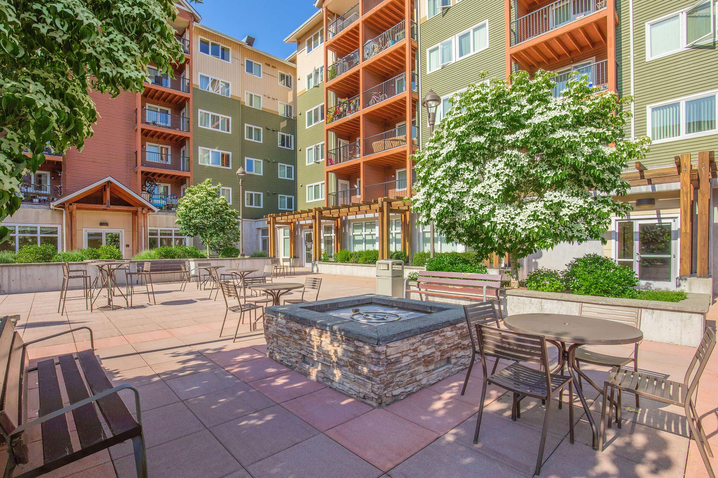 A bright outdoor courtyard area featuring landscaped greenery, seating arrangements with metal tables and chairs, and a fire pit. Surrounding buildings have colorful exteriors with balconies. Clear blue skies add to the inviting atmosphere, perfect for relaxation or social gatherings.