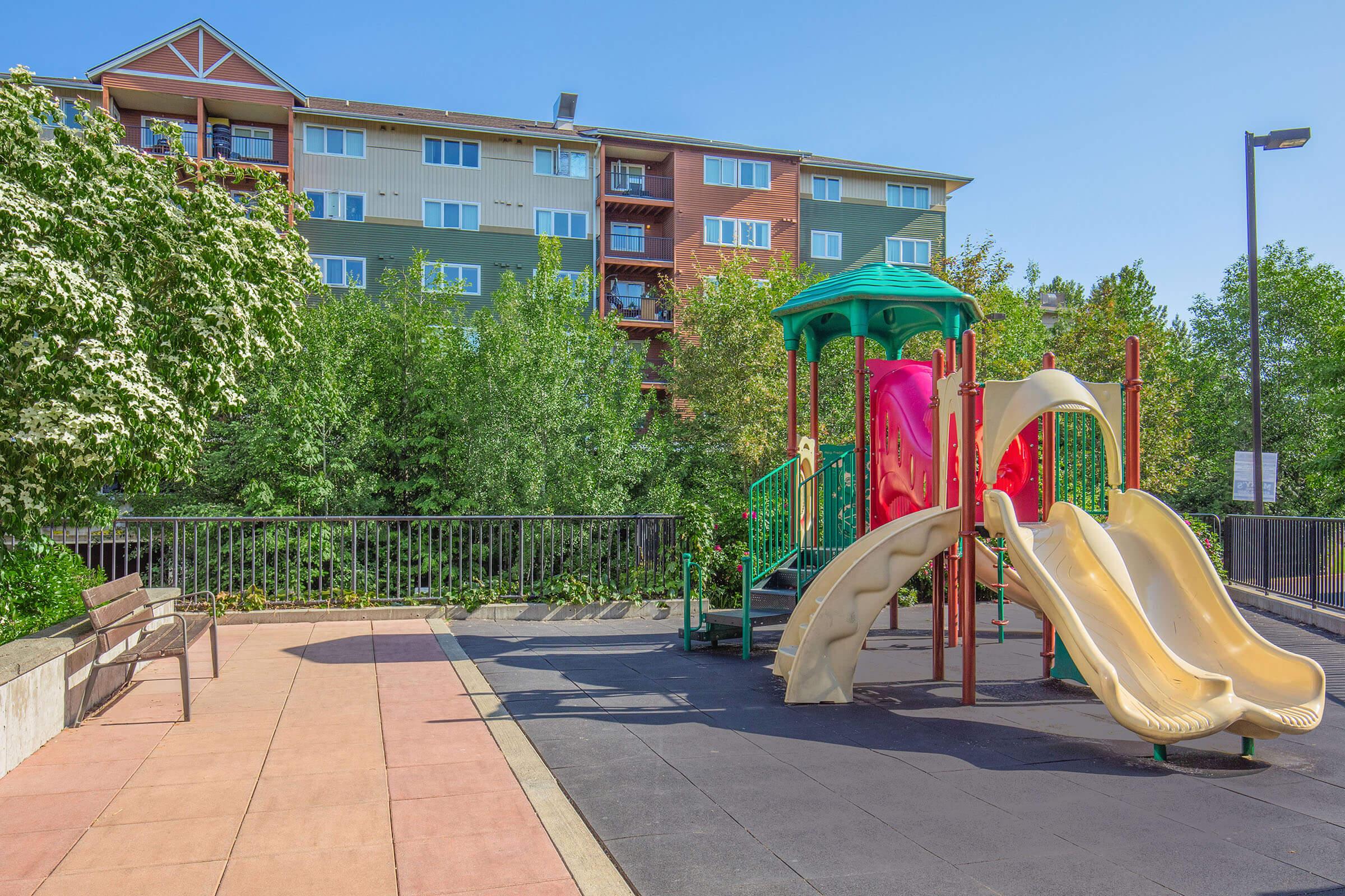 A colorful playground featuring a structure with two slides, surrounded by greenery and a paved area. In the background, an apartment building with multiple balconies is visible under a clear blue sky. A bench is placed nearby, facing the playground.
