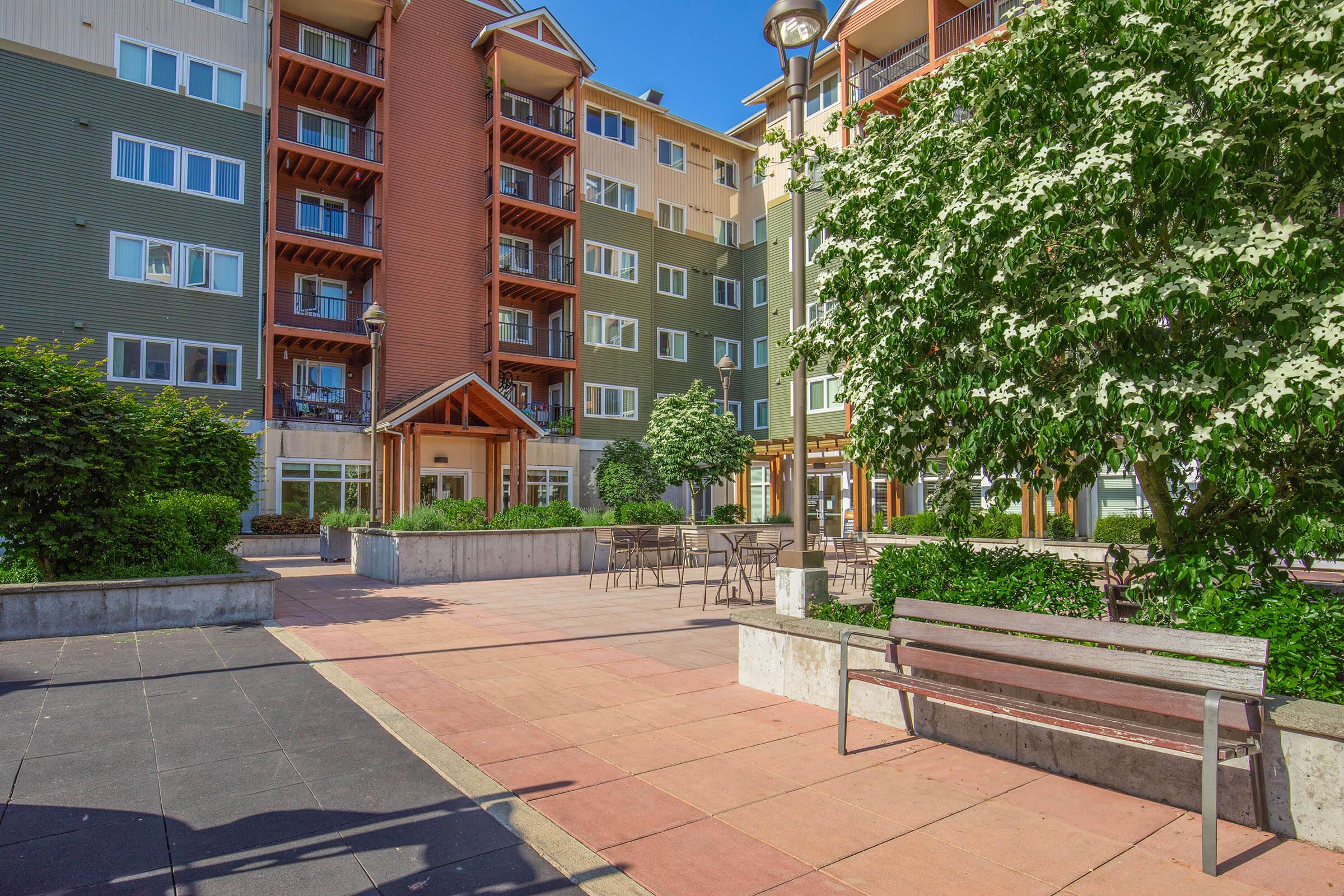 A view of a modern apartment complex with several multi-story buildings in the background. In the foreground, there is a paved area with benches, neatly landscaped greenery, and a few outdoor tables. The scene is bright and sunny, contributing to a welcoming and vibrant atmosphere.