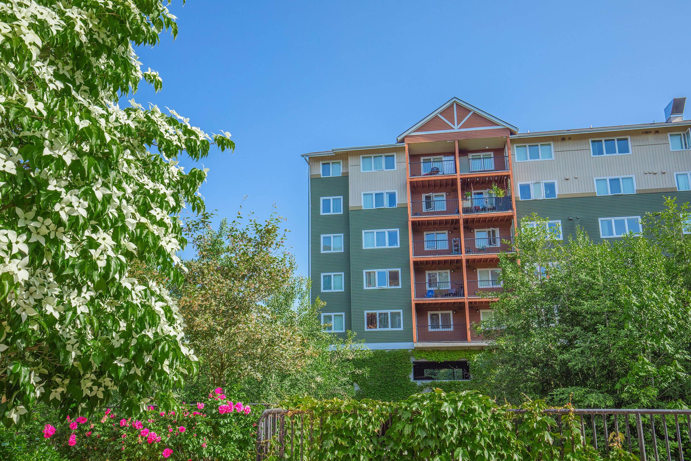 A multi-story building with green and beige siding, featuring balconies and large windows. In the foreground, blooming flowers and lush greenery frame the scene under a clear blue sky.