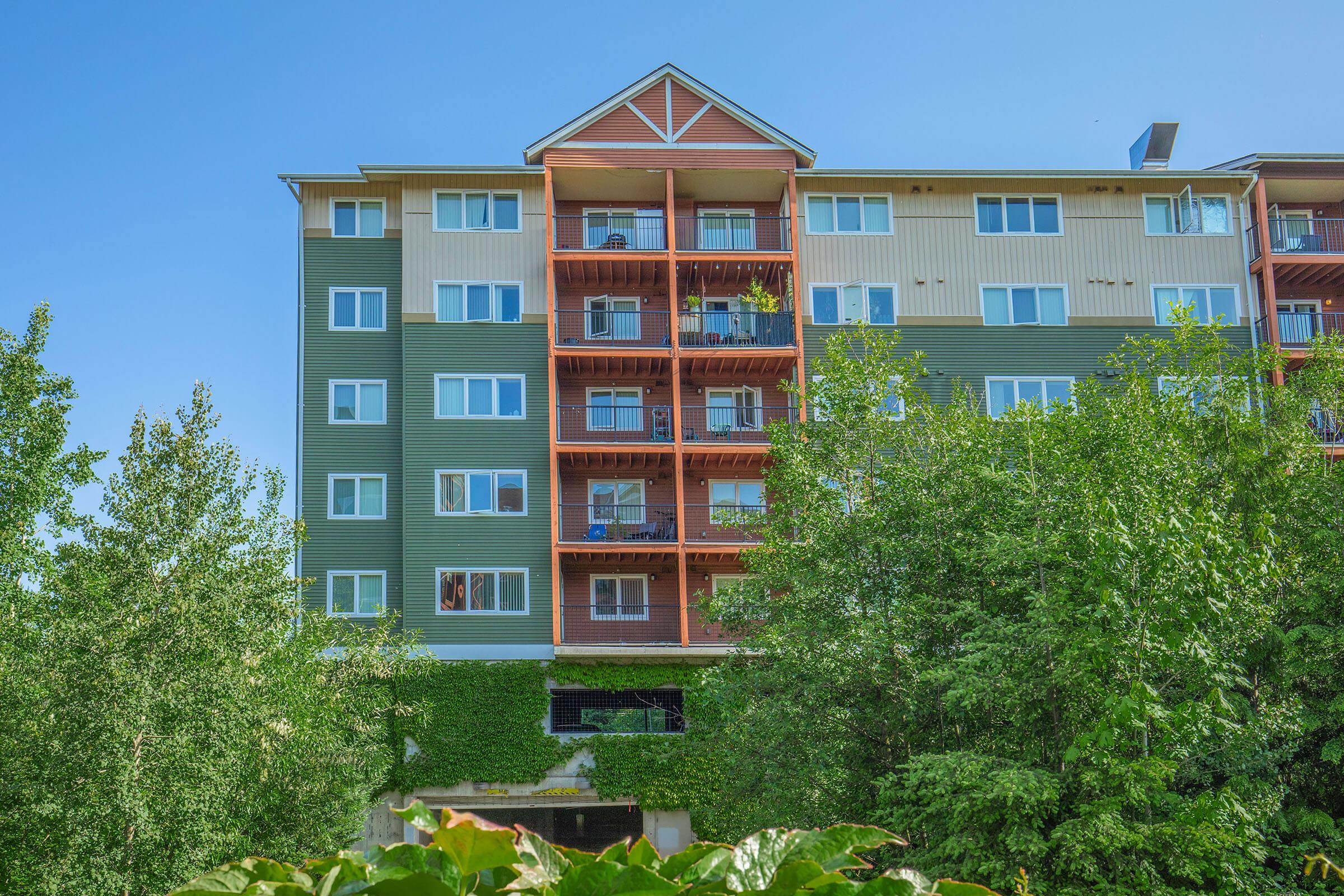 A multi-story residential building with green and tan siding. The upper floors feature balconies with plants, while the ground level is partially obscured by greenery. Clear blue sky above.