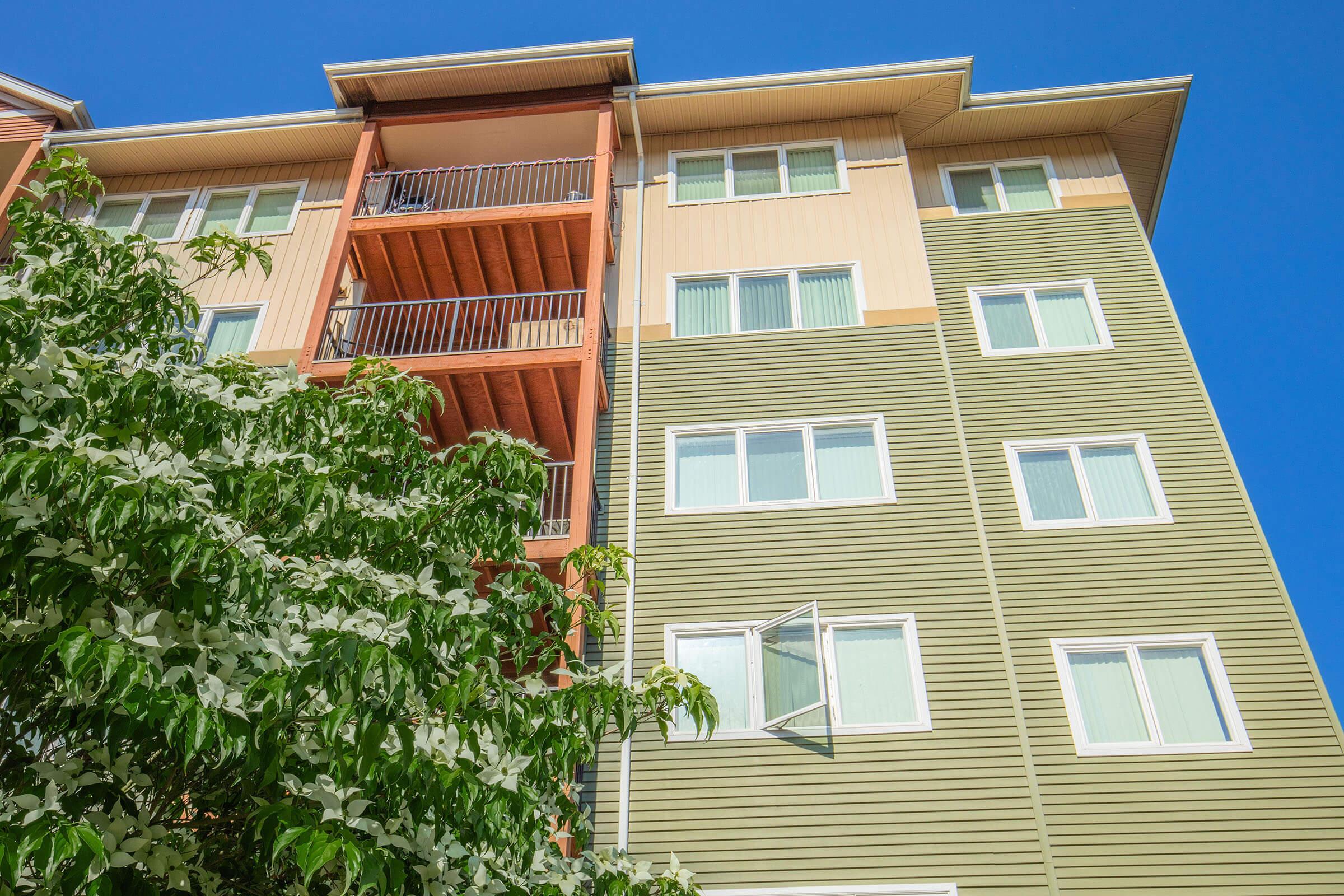 A modern multi-story apartment building featuring a mix of beige and green siding. There are multiple windows with light-colored frames, and one window is open. The foreground includes a lush green tree, providing a contrast to the building's facade against a clear blue sky.