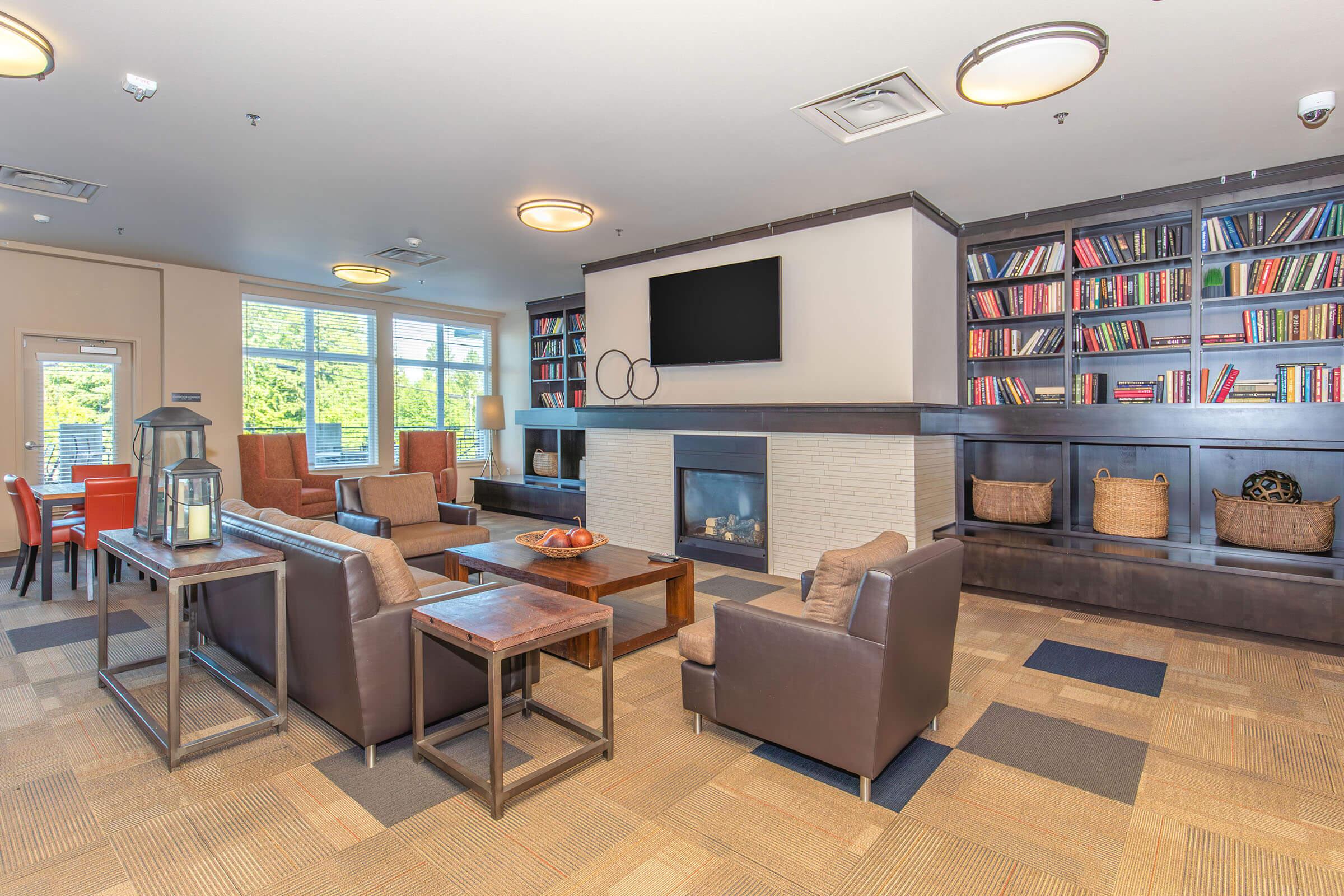 Cozy lounge area featuring comfortable brown leather seating, a wooden coffee table, and a modern fireplace. Shelves filled with books are displayed on one wall, while large windows let in natural light. Decorative elements like a lantern and baskets enhance the inviting atmosphere.