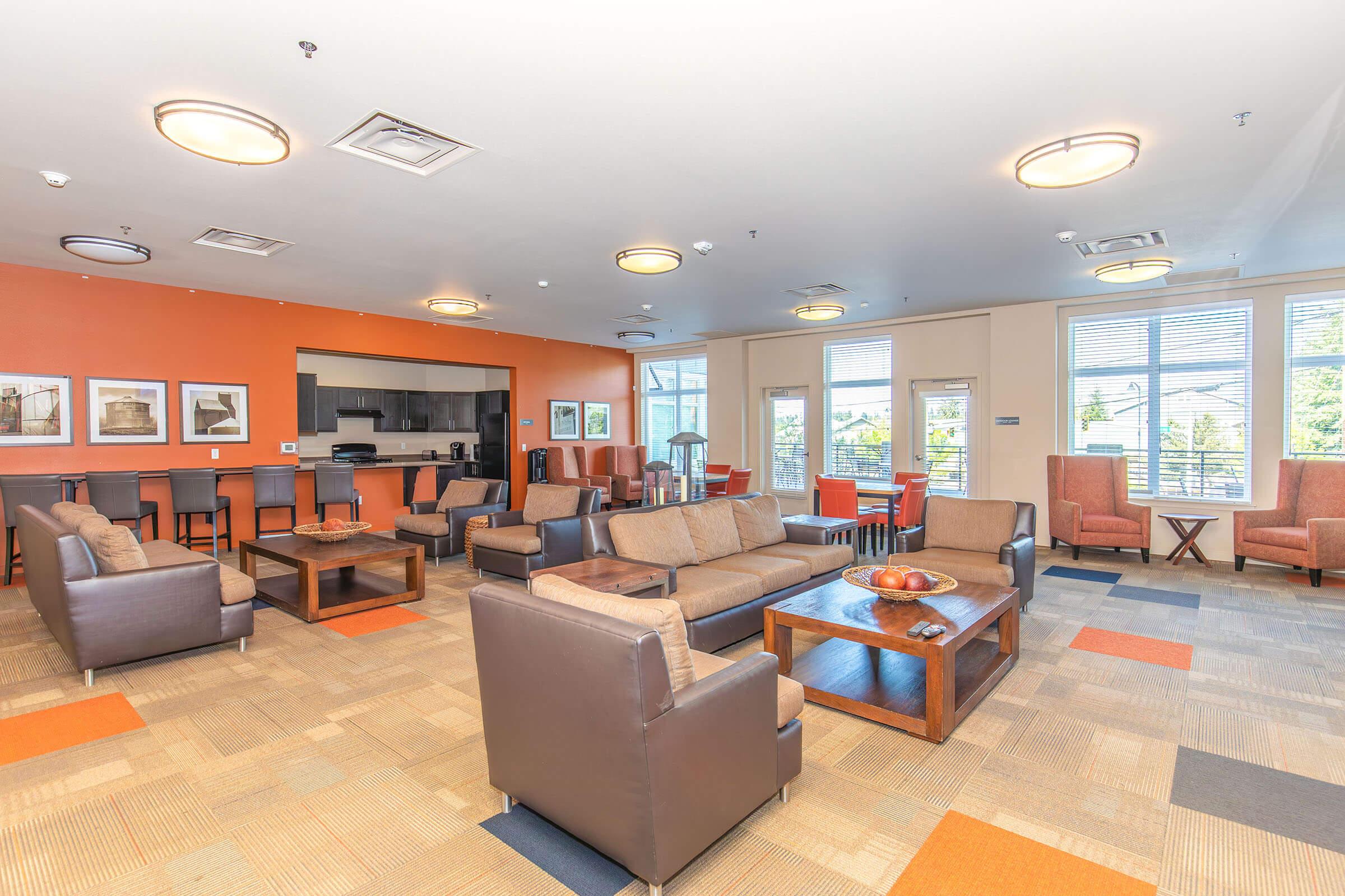A spacious lounge area featuring brown leather sofas, armchairs, and a wooden coffee table. The walls have an orange accent, and there are framed artworks. A kitchen area is visible in the background, along with barstool seating. Natural light streams in through large windows, creating a welcoming atmosphere.