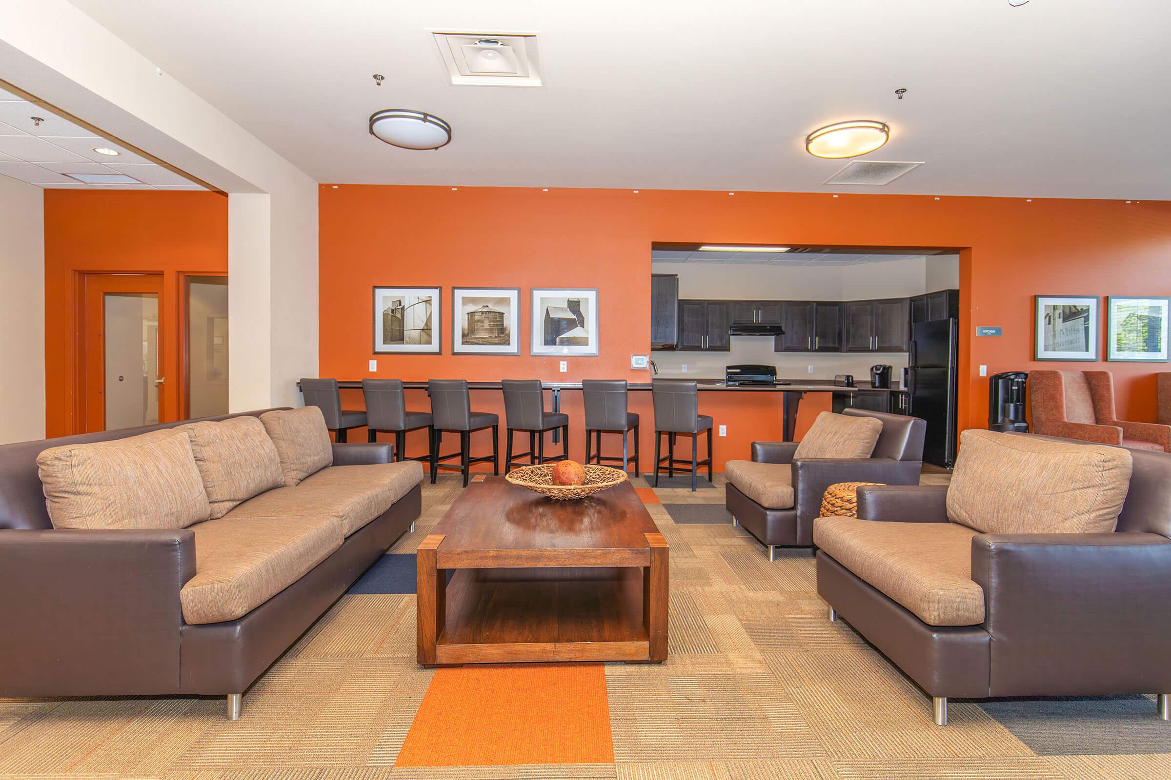 A modern lounge area featuring two brown leather armchairs and a large brown wooden coffee table with a decorative bowl. In the background, there are bar-style stools at a counter, a kitchen area, and framed black-and-white photographs on the orange accent wall, creating a cozy and inviting atmosphere.
