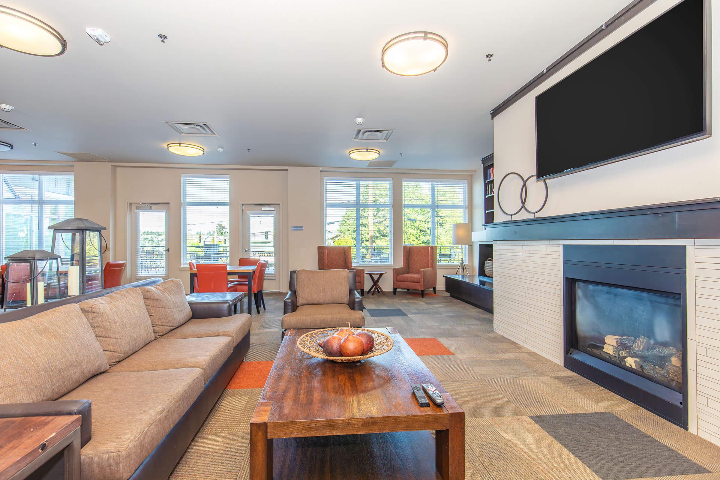 A modern lounge area featuring a brown sofa and a wooden coffee table with a decorative bowl. There's a large flat-screen TV mounted on the wall above a fireplace. Bright windows provide natural light, and the space includes a seating area with chairs and a dining table in the background.