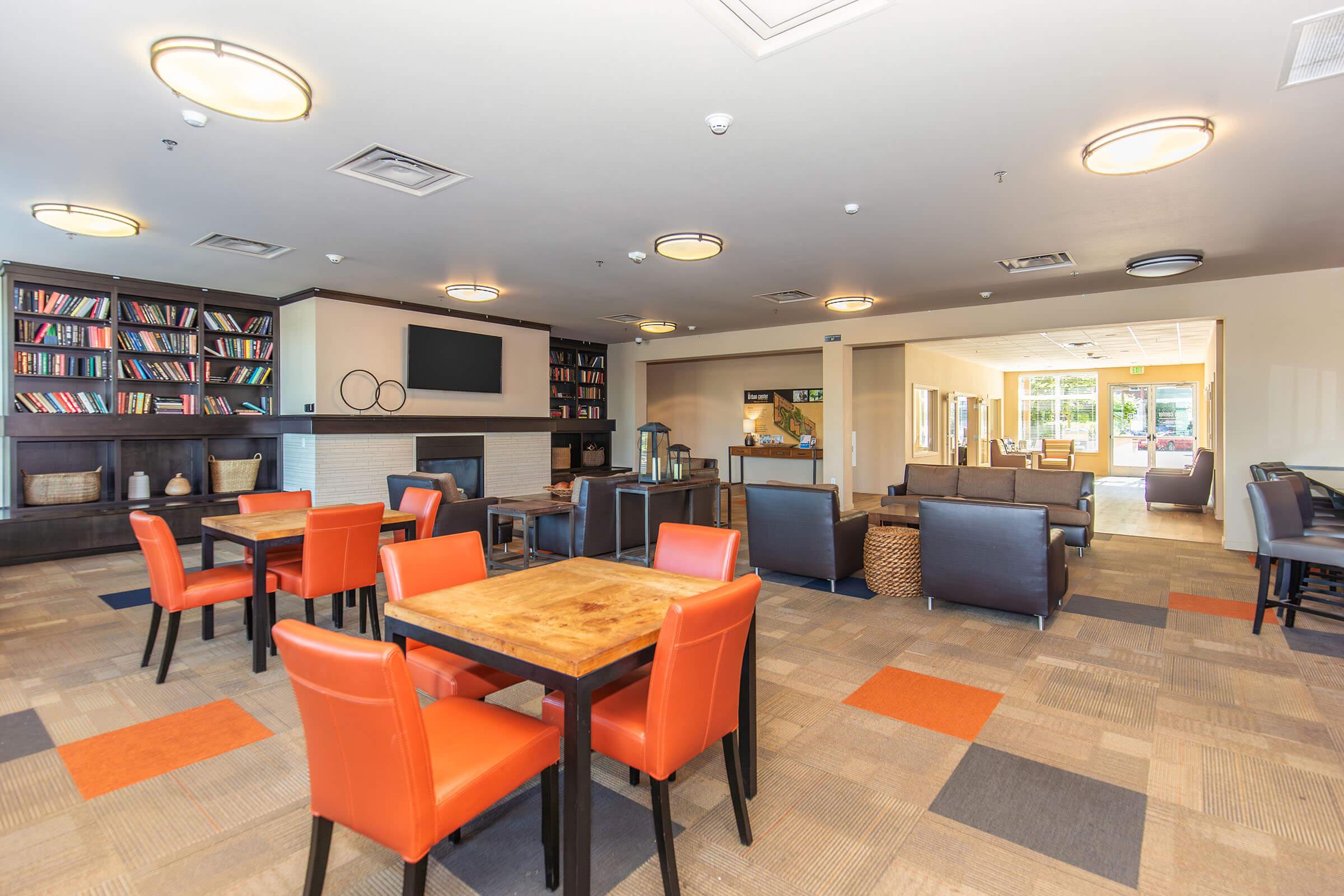 A modern lounge area featuring comfortable seating with orange and black chairs, wooden tables, a bookshelf filled with books, and large windows providing natural light. The space is designed for relaxation and social interaction, with a fireplace and decorative elements enhancing the atmosphere.