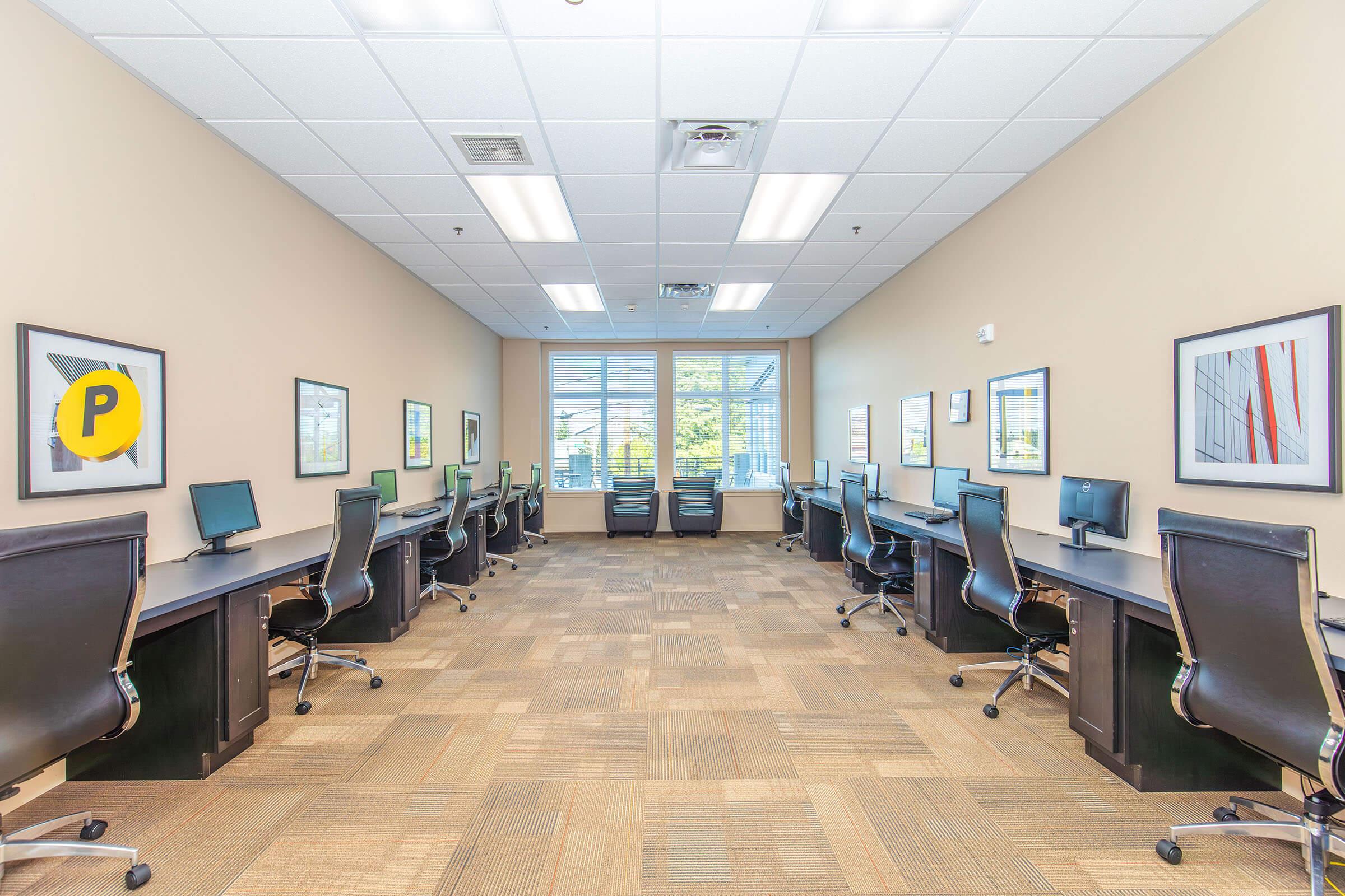 A spacious office area featuring several computer stations with ergonomic chairs. The room has beige walls and large windows providing natural light. Framed artwork adorns the walls, adding a professional touch. Two chairs are positioned by the windows, suggesting a collaborative space.