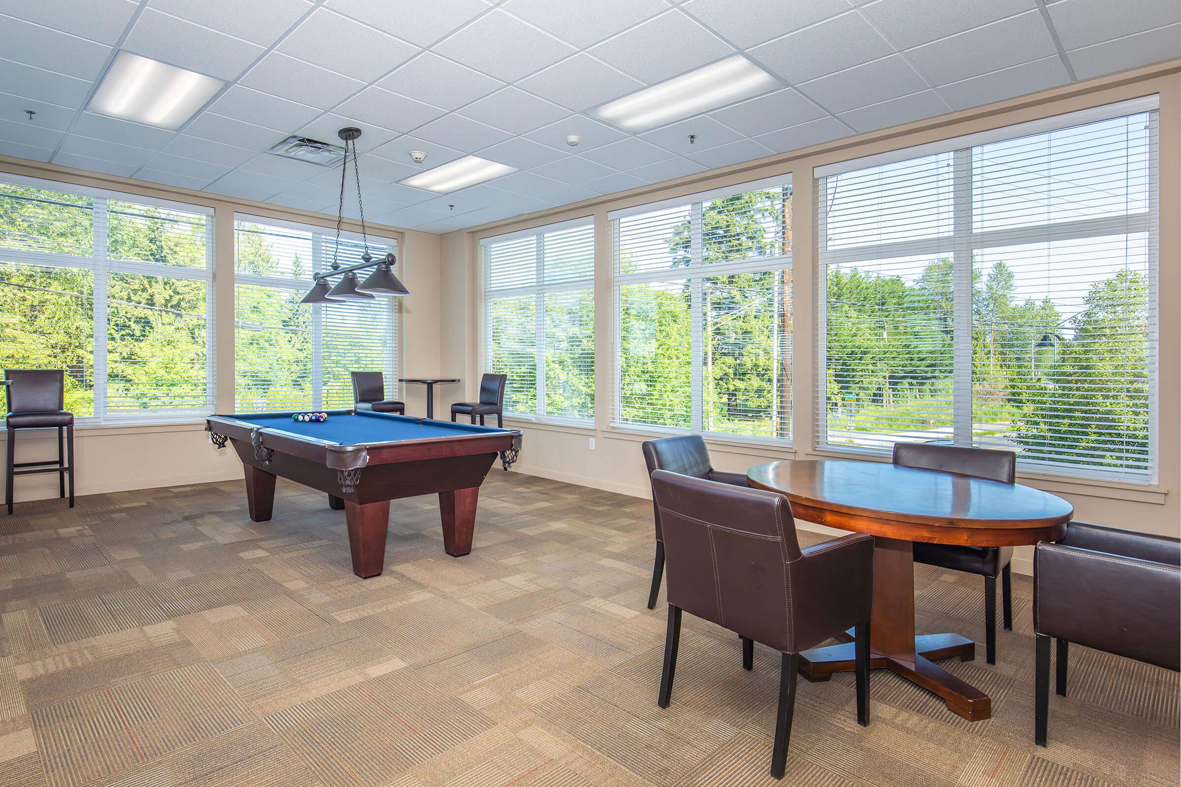 A spacious indoor recreation room featuring a pool table, a round wooden dining table, and several dark leather chairs. Large windows let in natural light and provide a view of greenery outside, creating a bright and inviting atmosphere. The flooring is carpeted with a neutral pattern.