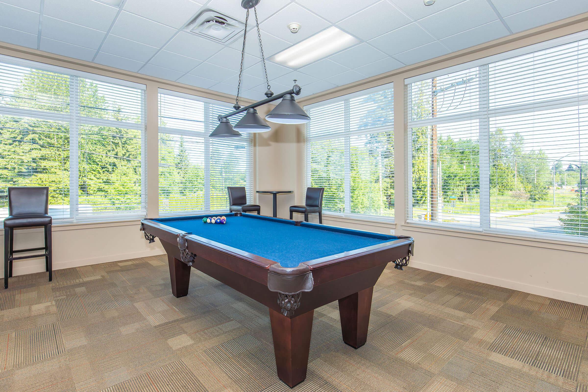 A well-lit game room featuring a blue pool table with a set of billiard balls arranged on it. Large windows provide natural light, and there are two bar stools in the background. The flooring is a light brown carpet, and the walls are painted in a neutral tone, creating a welcoming atmosphere.