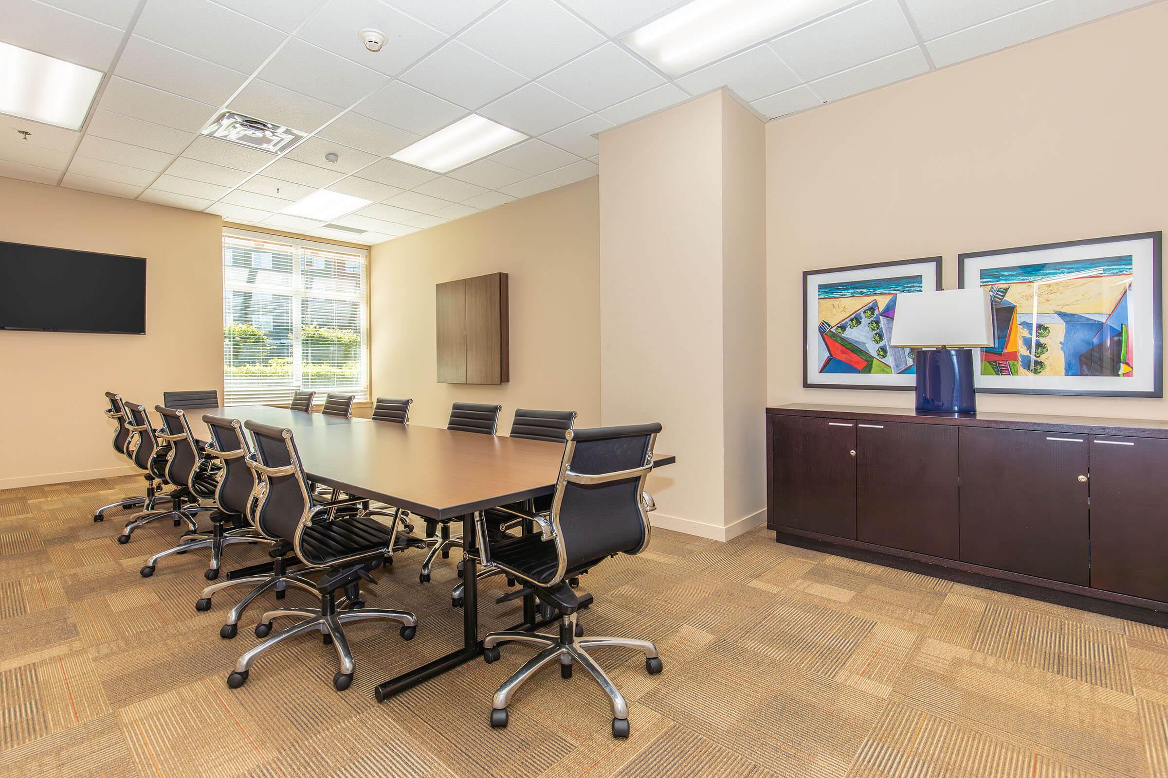 A modern conference room featuring a large wooden table surrounded by black office chairs. A wall-mounted television is displayed on one side, while on the opposite wall, two framed artwork pieces are visible above a dark wood cabinet. The room is well-lit with large windows allowing natural light in.