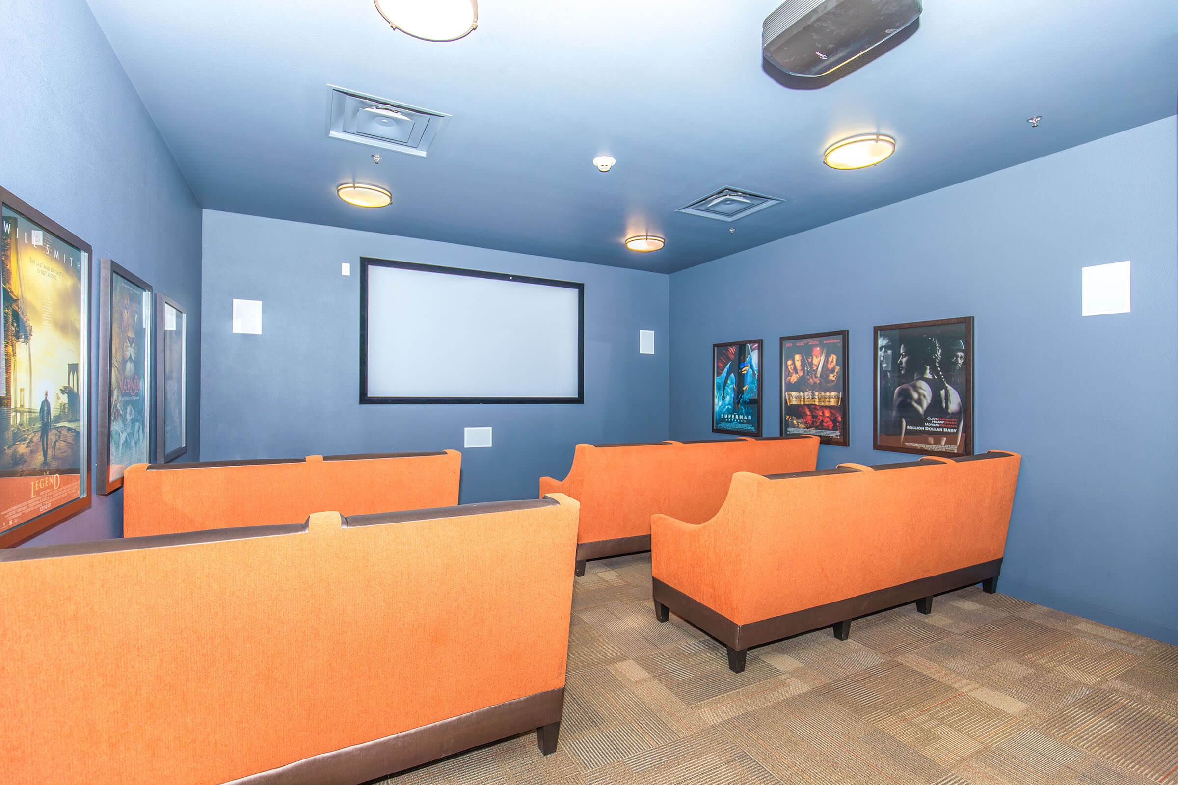 A small home theater room with dark blue walls and orange seating. The room features a large screen on one wall, surrounded by framed movie posters. The carpet is light-colored, and overhead lights provide illumination. The layout suggests a cozy atmosphere for watching films.