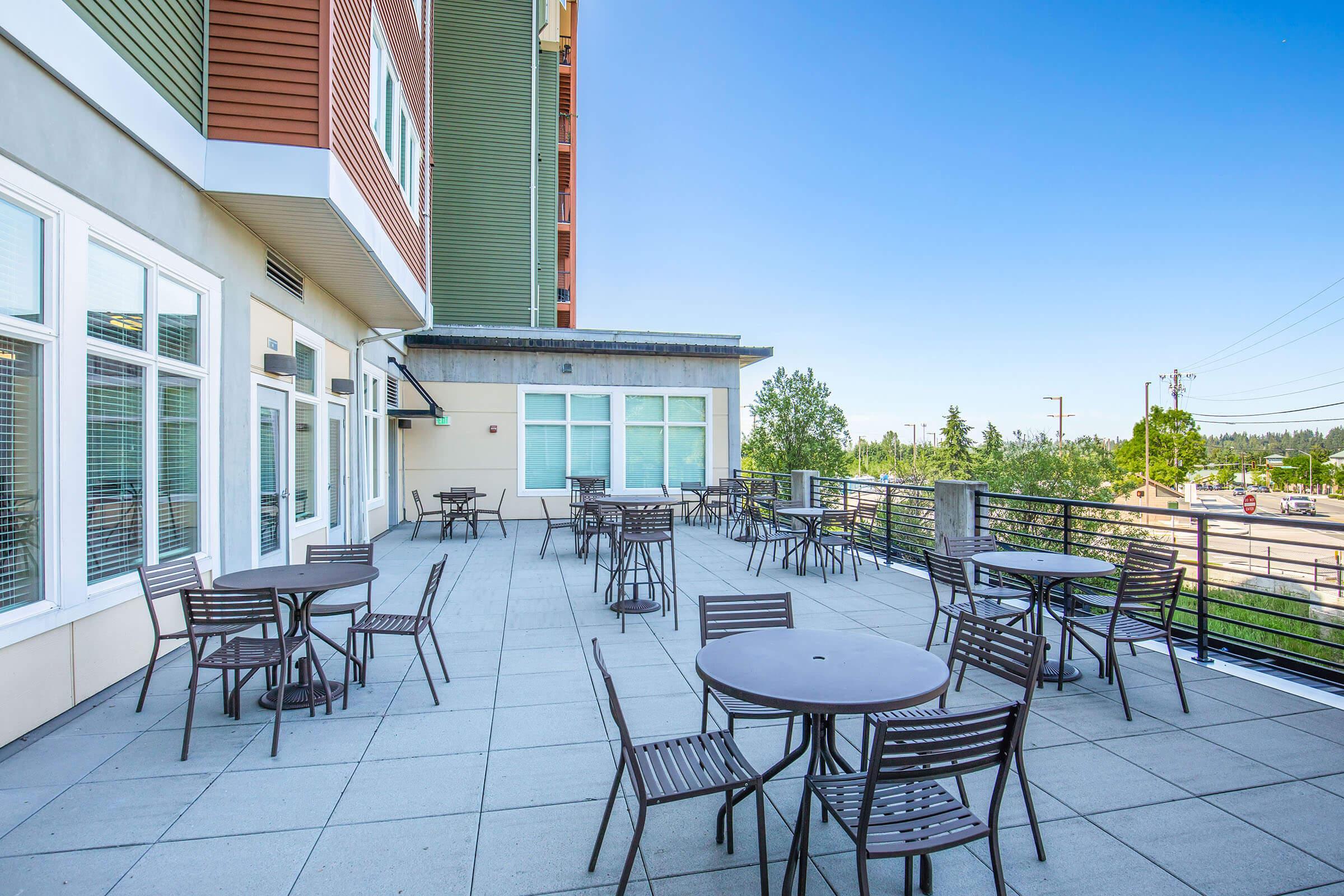 Outdoor patio area featuring several round metal tables and chairs, surrounded by trees and a clear blue sky. The setting includes a building with a modern architectural design and large windows, providing a spacious and inviting atmosphere for dining or socializing.
