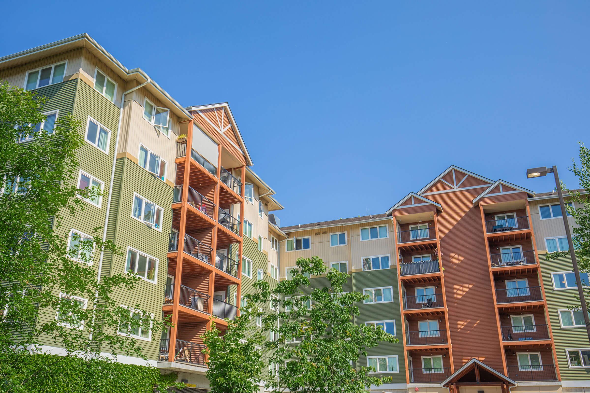 A multi-story residential building with a mix of green, beige, and orange siding. The structure features balconies and large windows, surrounded by trees under a bright blue sky. The architectural design includes gabled roofs and a modern aesthetic.