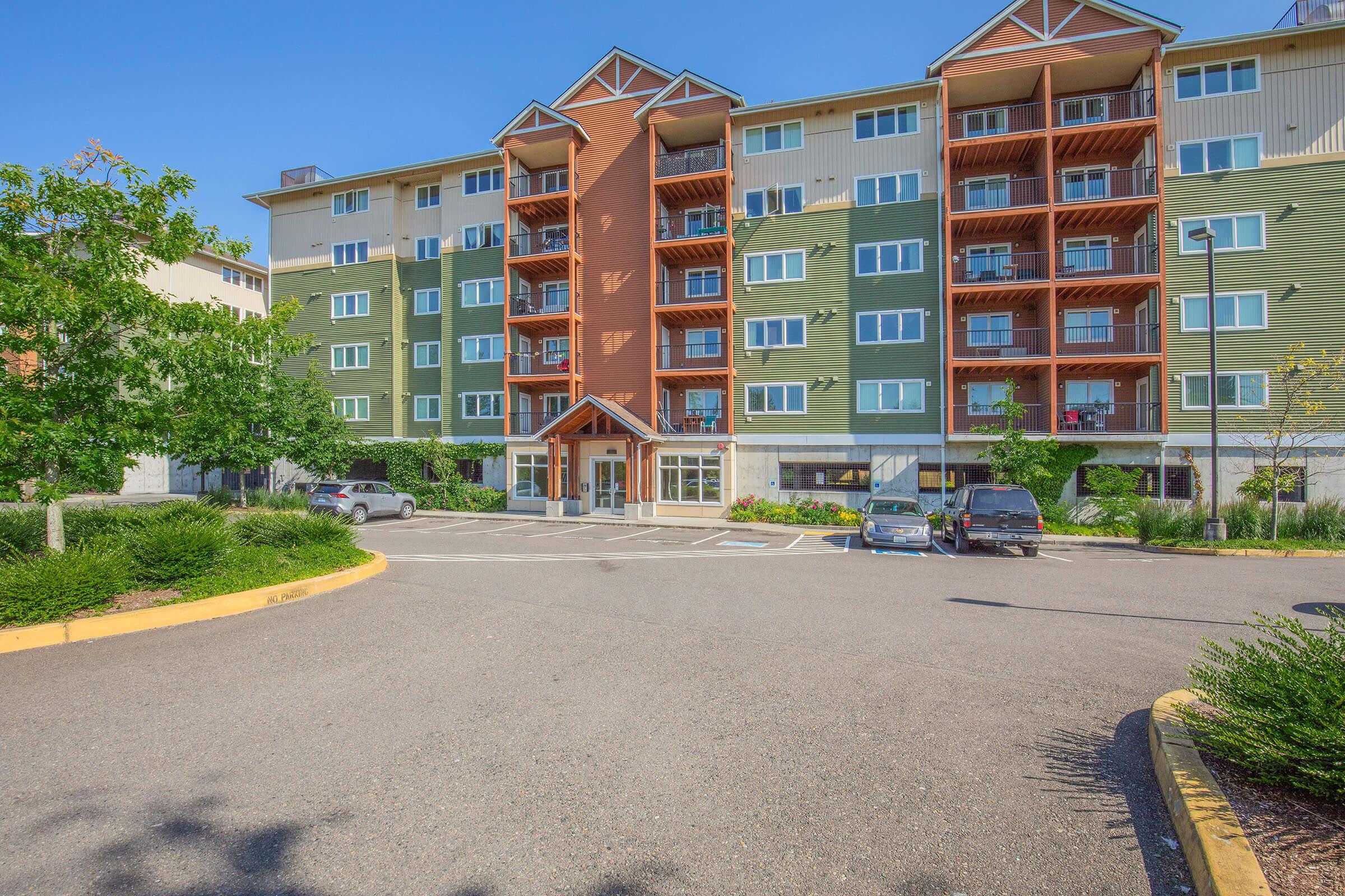 A four-story apartment building with a combination of green and orange exterior features. The front entrance is highlighted, and there is a small parking area with several cars. Surrounding landscaping includes trees and shrubs, under a clear blue sky.